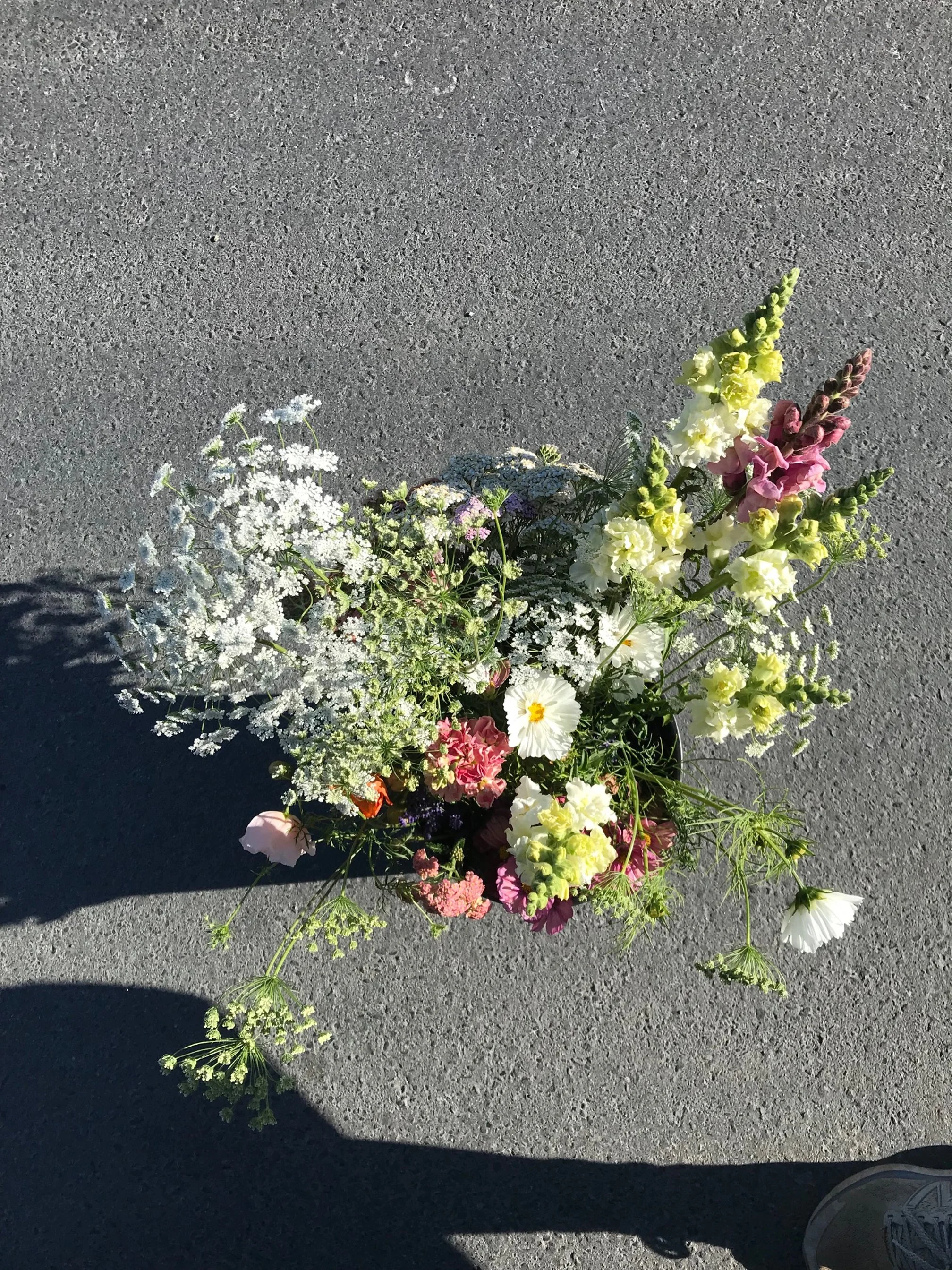 A colorful bouquet of various flowers laying on gray pavement, with a shadow and a visible sneaker in the bottom right corner.