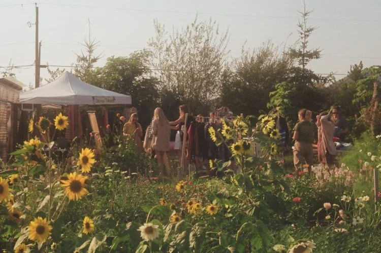 People shopping at an outdoor market or yard sale with sunflowers and other flowers in foreground, tents, and trees in background.