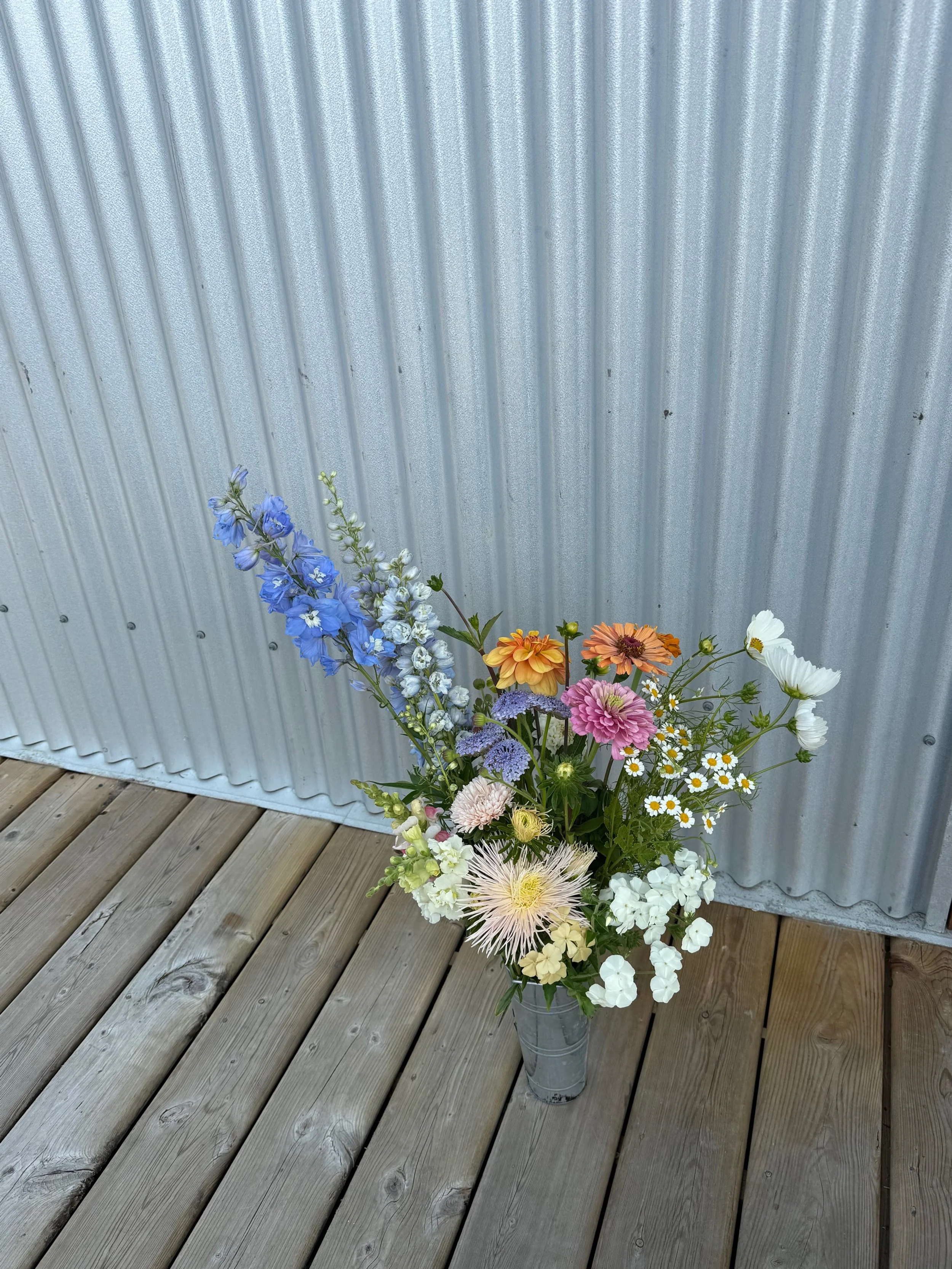 Colorful bouquet of various flowers in a metal bucket on wooden deck against a corrugated metal wall.