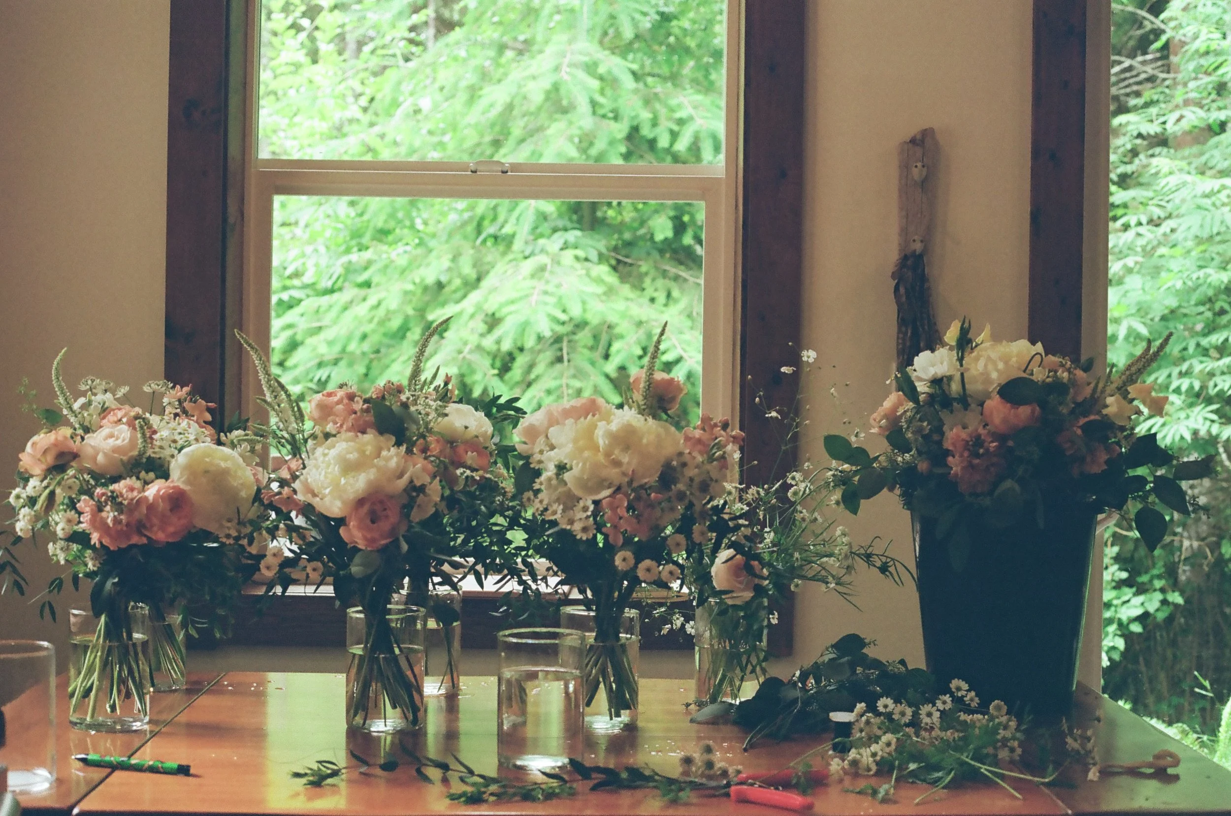 A table with several glass vases filled with pink and white flowers, next to a large black pot with similar flowers, on a wooden table in front of a window showing green trees outside.
