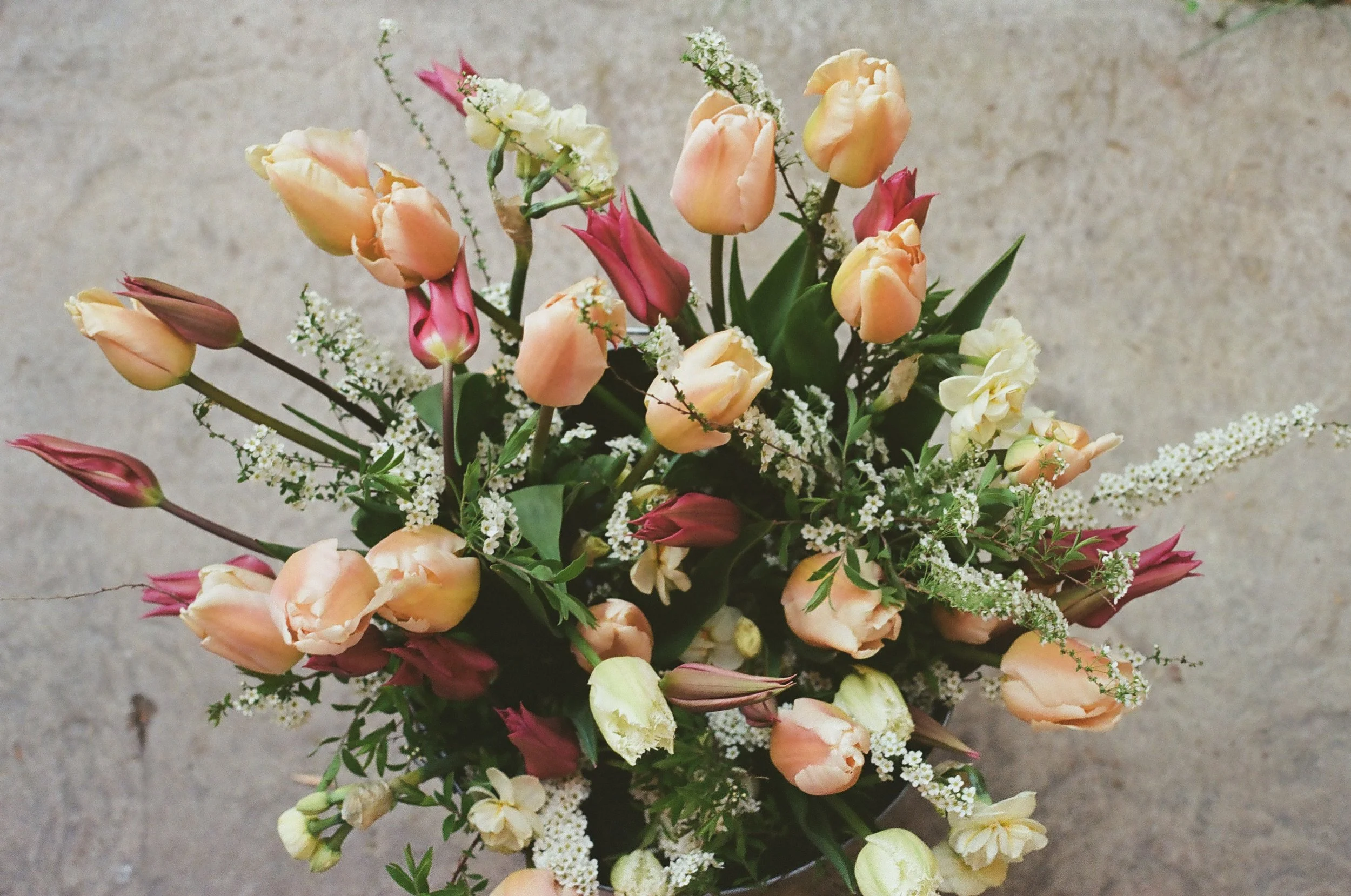 A bouquet of peach, pink, and cream-colored tulips with greenery and small white filler flowers, placed on a gray surface.