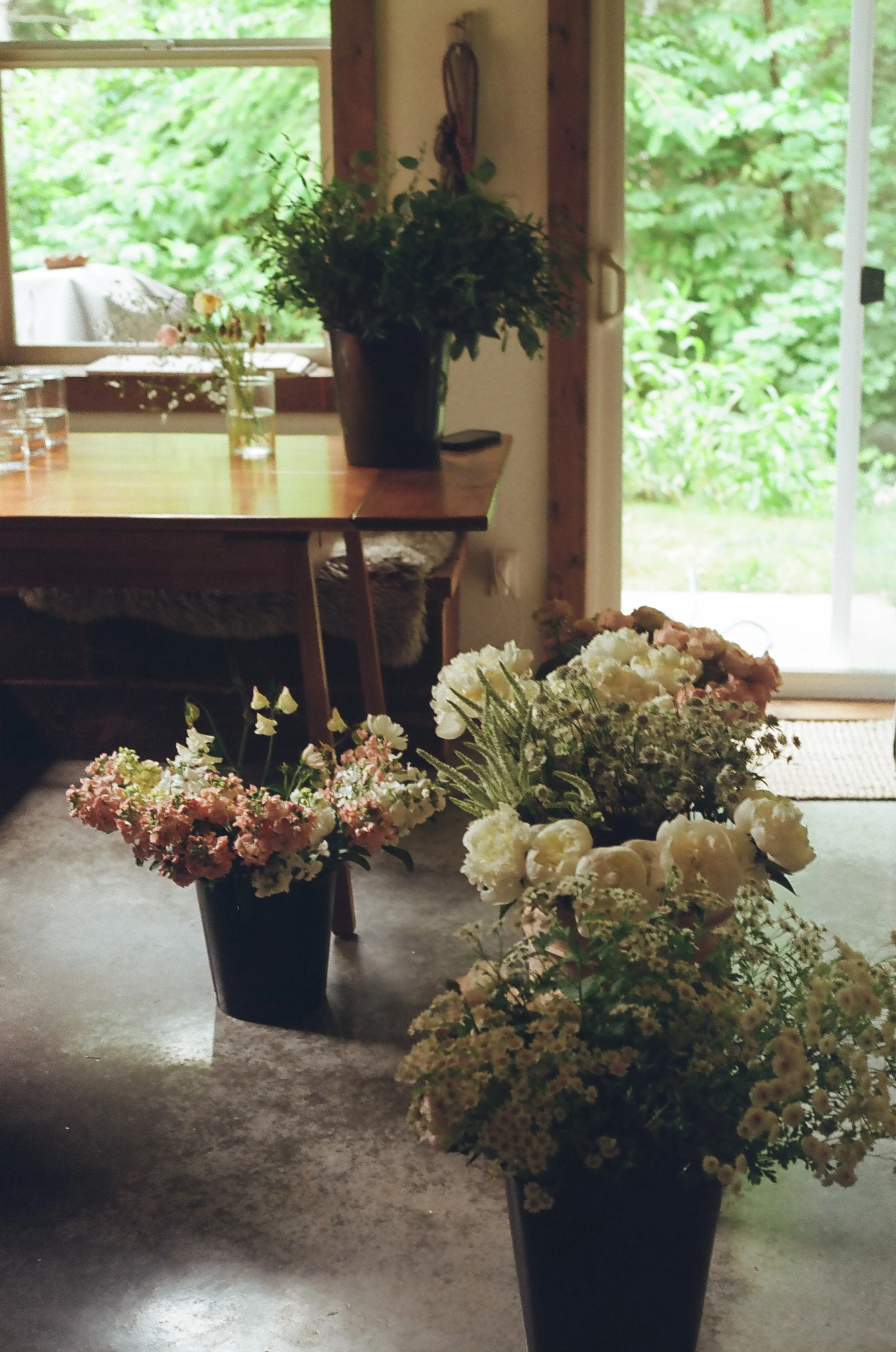 A room with a wooden table and flowers in buckets, with a window revealing green foliage outside.