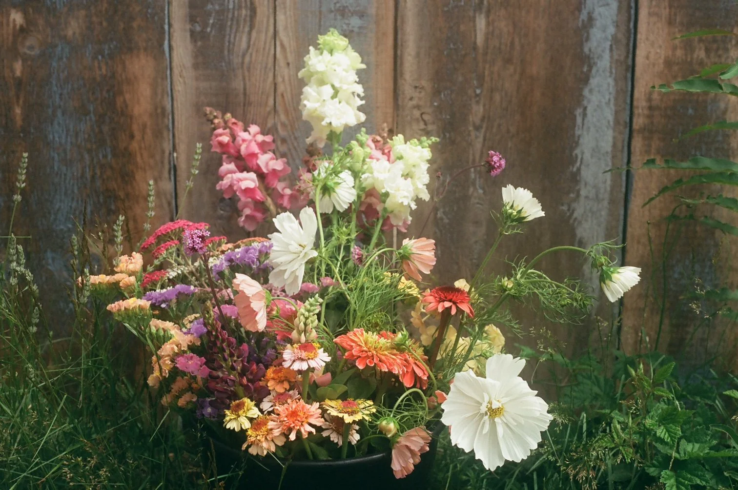 A vase filled with colorful flowers including white, pink, orange, purple, and red blooms, set against a wooden background.