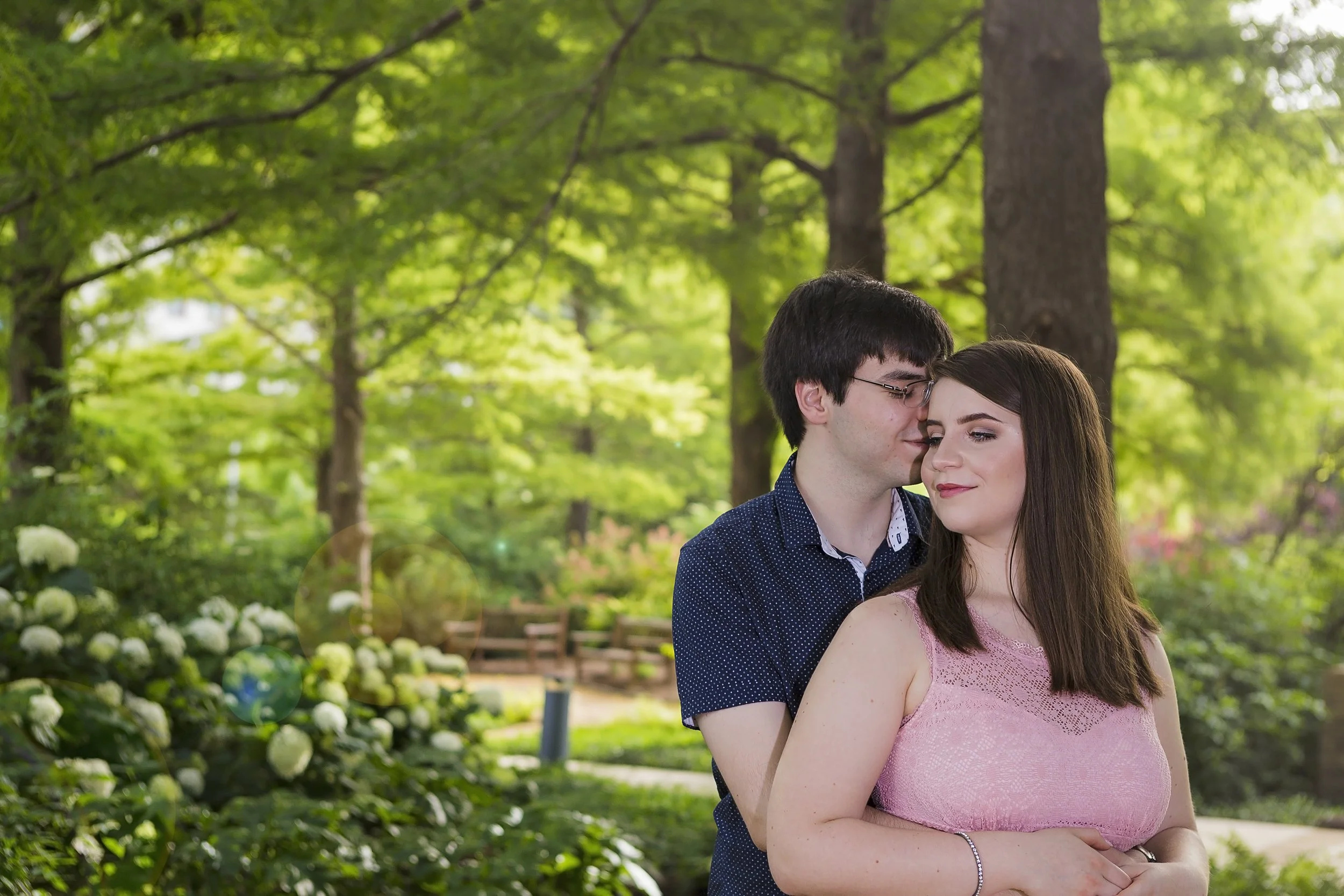 Outdoor engagement session of  couple embracing in a park with green trees and bushes in the background.