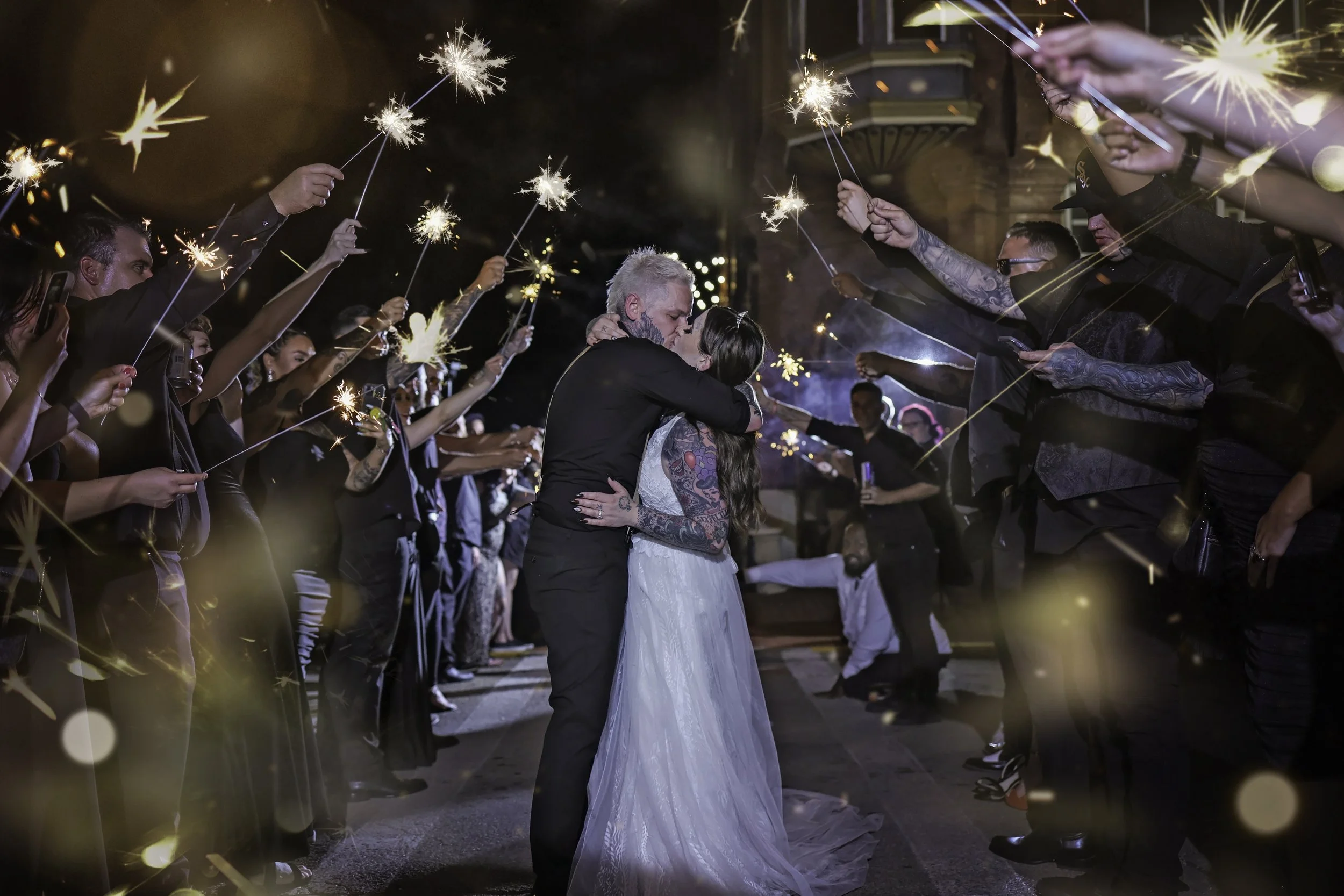 A couple sharing a kiss during their wedding surrounded by friends holding sparklers at night.