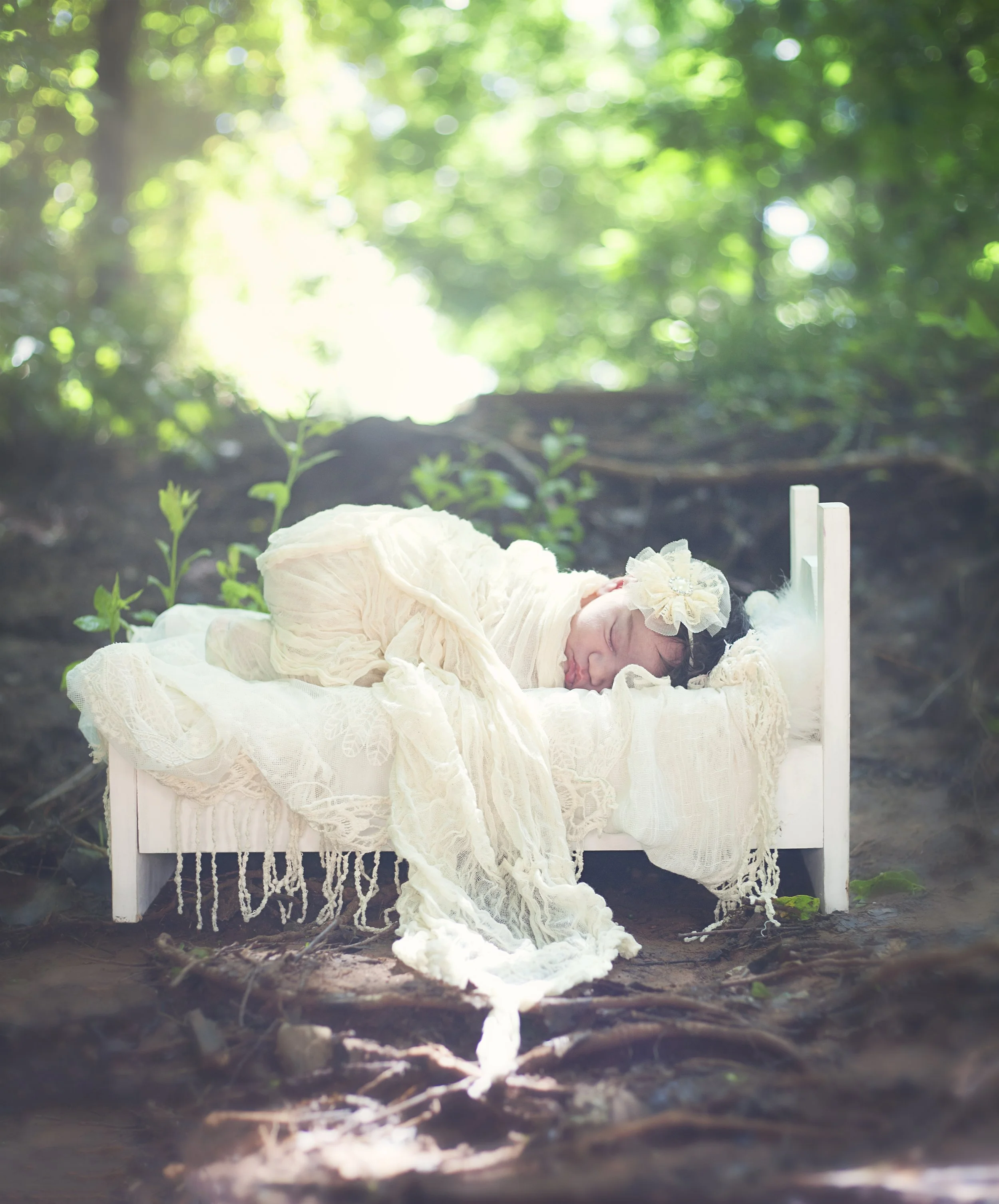 A baby girl sleeping on a white miniature bed outdoors in a forest setting with sunlight filtering through the trees.