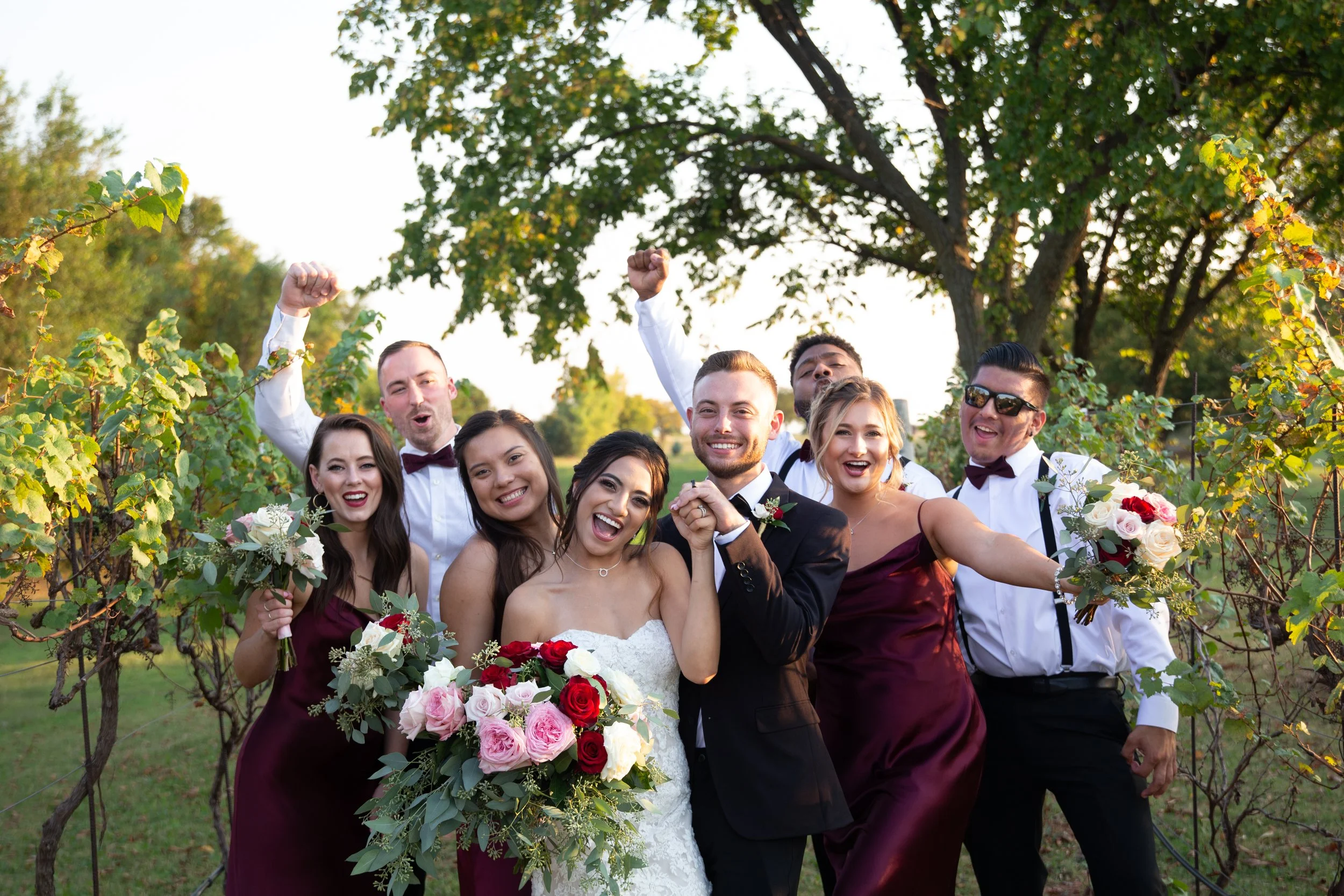 A group of wedding celebrants, including a bride in a white dress, a groom in a black suit, bridesmaids in burgundy dresses, and groomsmen in white shirts with burgundy bow ties, celebrating outdoors with smiles, bouquets, and expressive gestures und