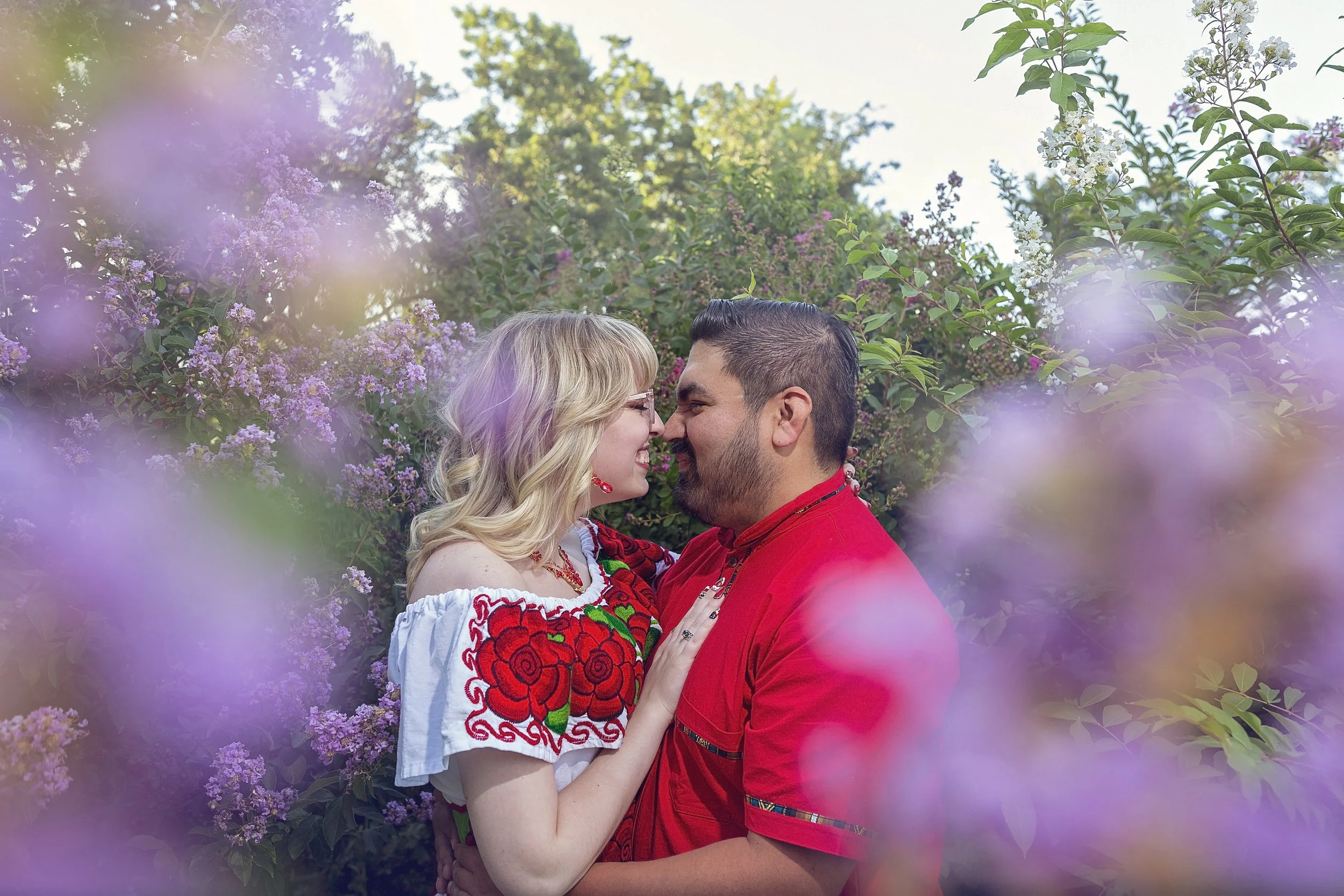 Engagement session touching noses, surrounded by pink and white blooming flowers and greenery in a garden, with the woman wearing traditional Mexican dress.