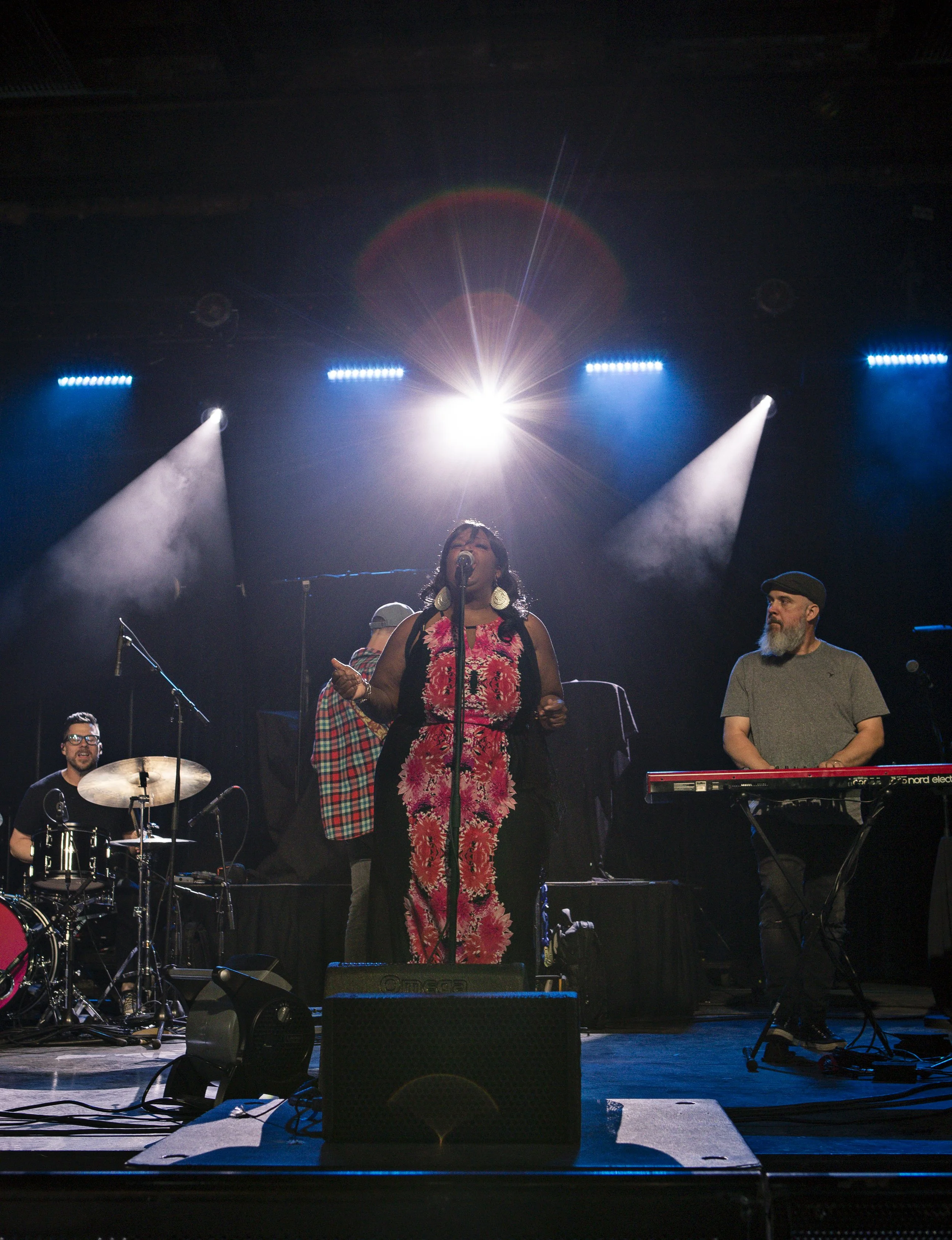 A woman singing on stage with a band, stage lights shining behind her, and a keyboardist and drummer accompanying her.