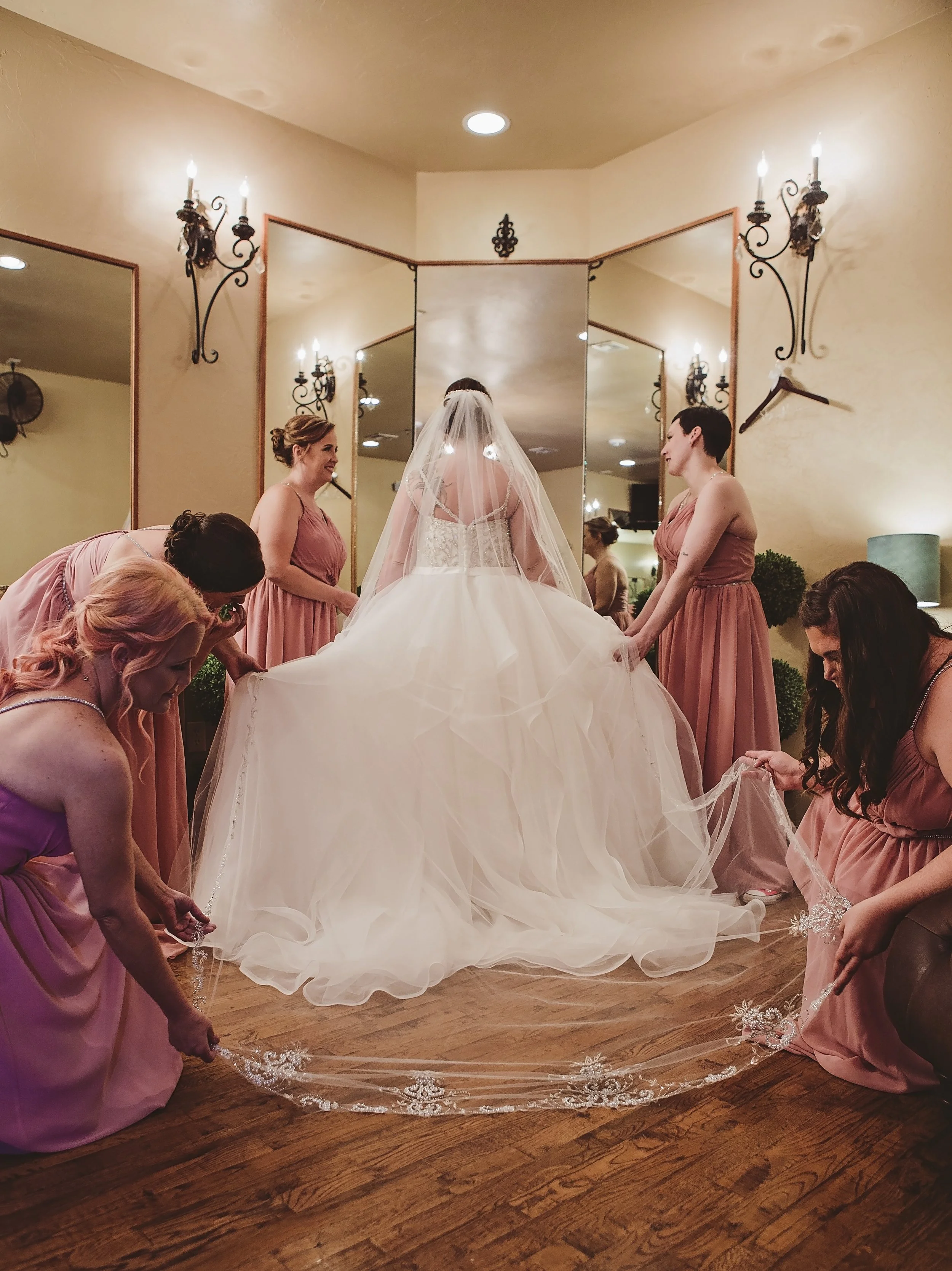 A bride in a wedding gown stands in front of a mirror, surrounded by bridesmaids in pink dresses adjusting her dress and veil in a warmly lit room.