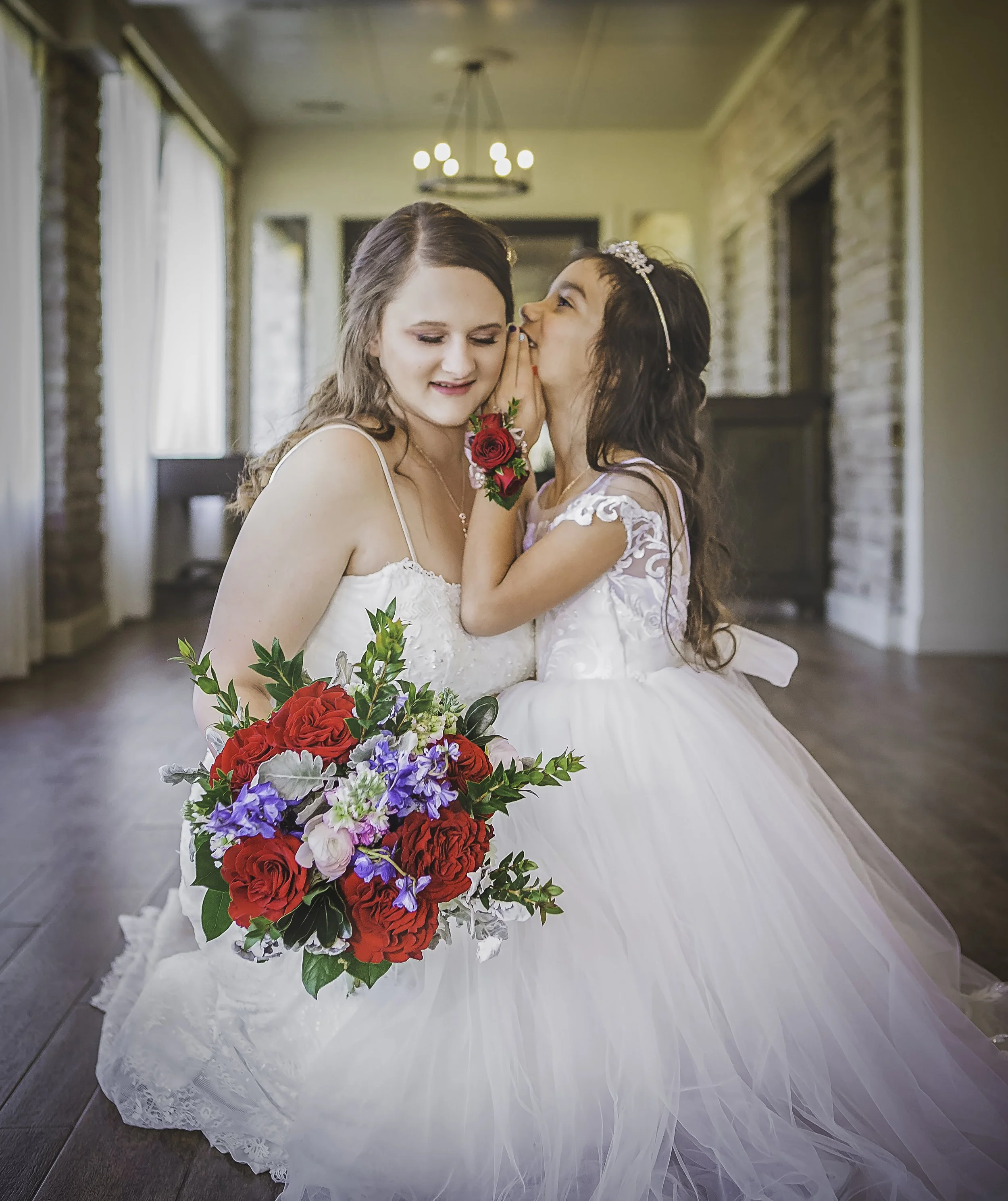 A bride and a young girl, likely a flower girl, sharing a special moment during a wedding. The bride is dressed in a white wedding gown and holding a vibrant bouquet of red, purple, and white flowers. The young girl is whispering to the bride, wearin