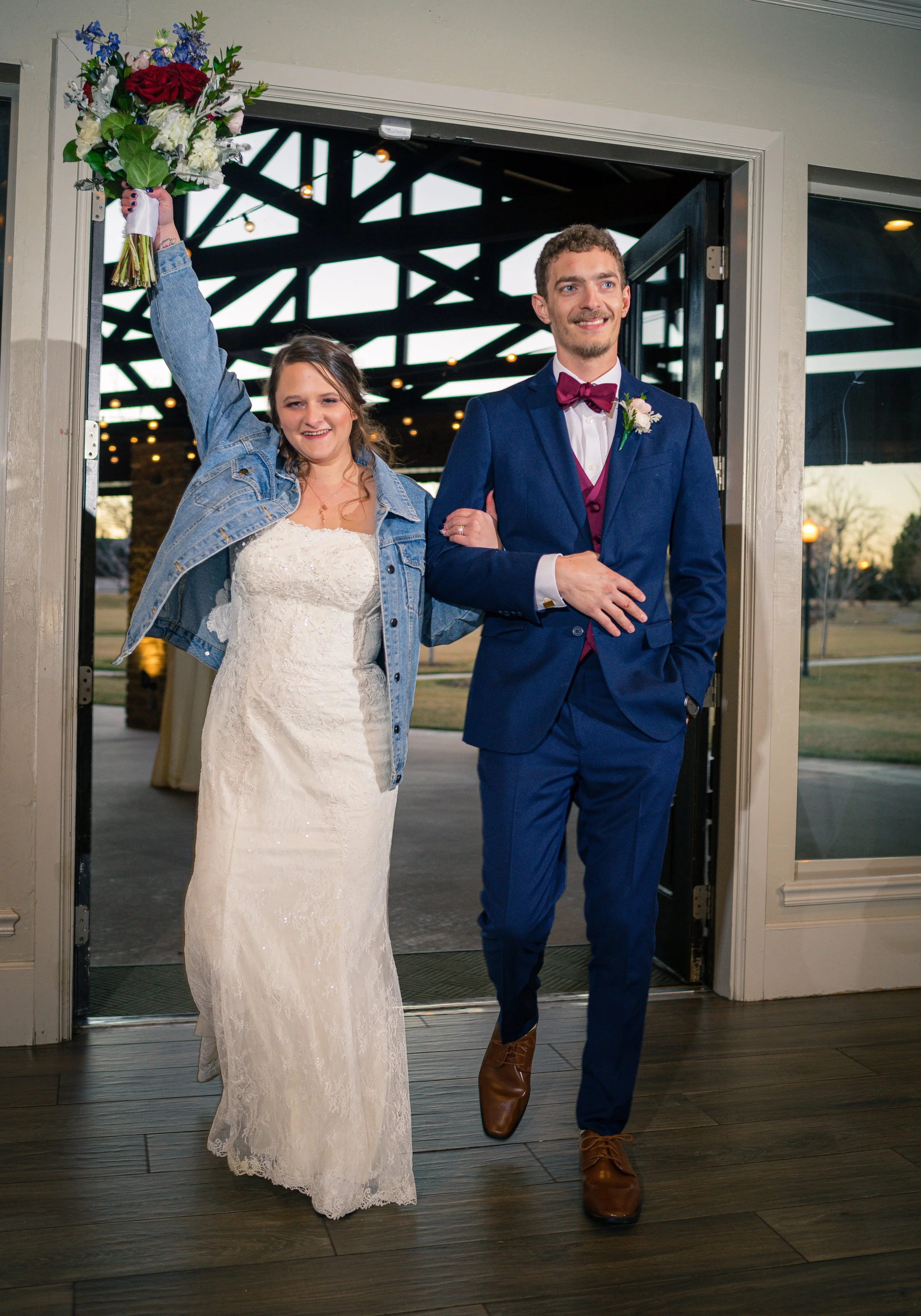 A bride and groom walk into a wedding reception, the bride holding a bouquet and wearing a denim jacket over her wedding dress, while the groom wears a blue suit with a burgundy bow tie, and they both smile