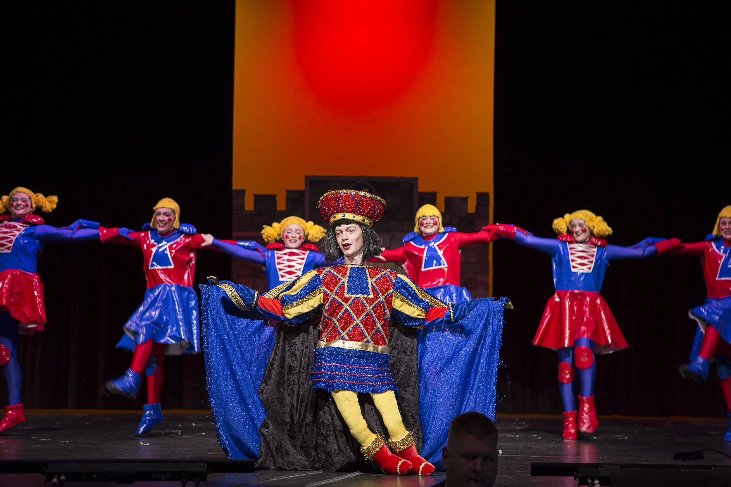 Theater performance with a woman in colorful costume center stage, surrounded by dancers in bright red, blue, and yellow outfits with yellow pigtails, on a stage with a backdrop resembling a castle and orange sky.