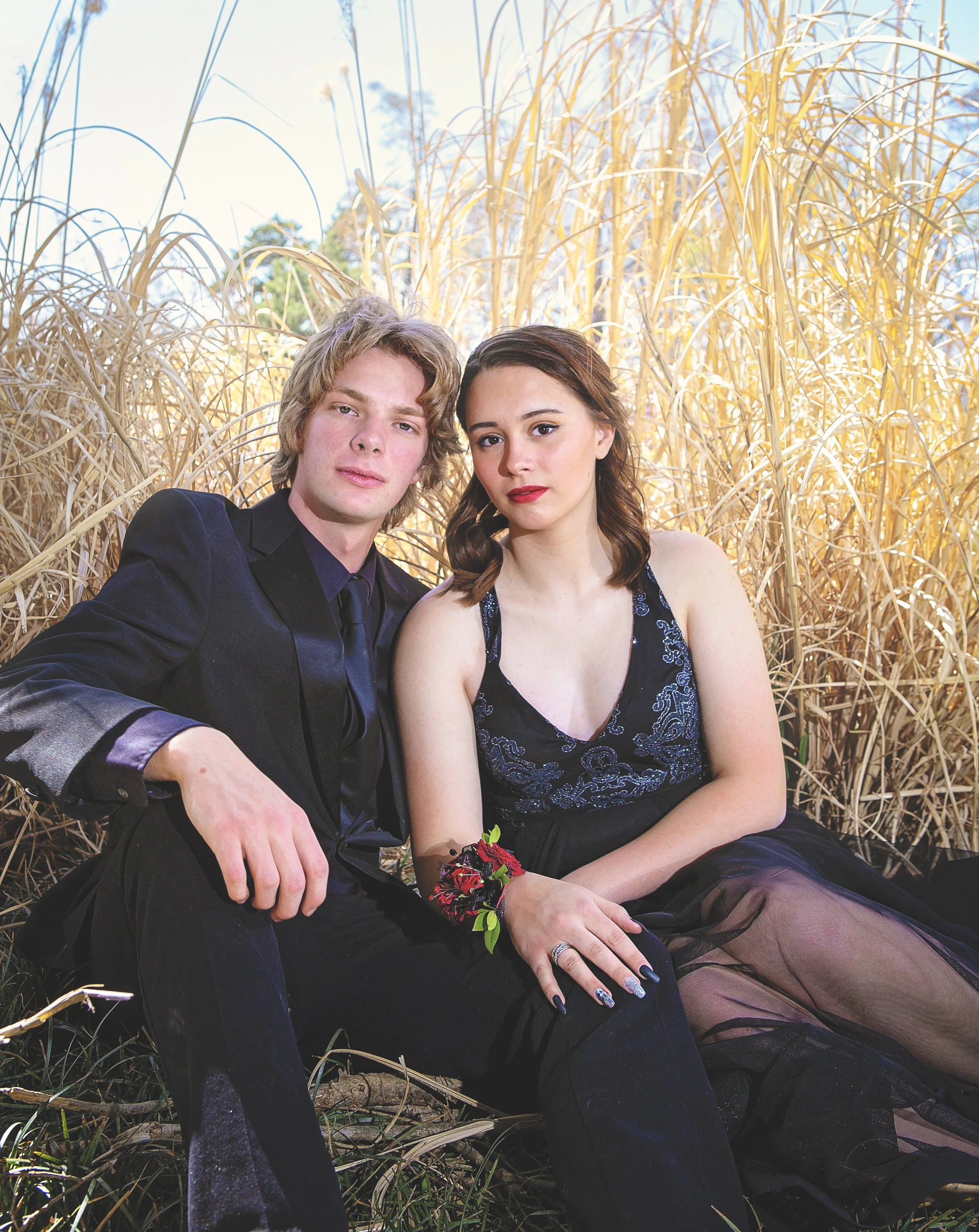 A young man and woman sitting in a field of tall, dry grass on a sunny day, dressed in formal attire with serious expressions.