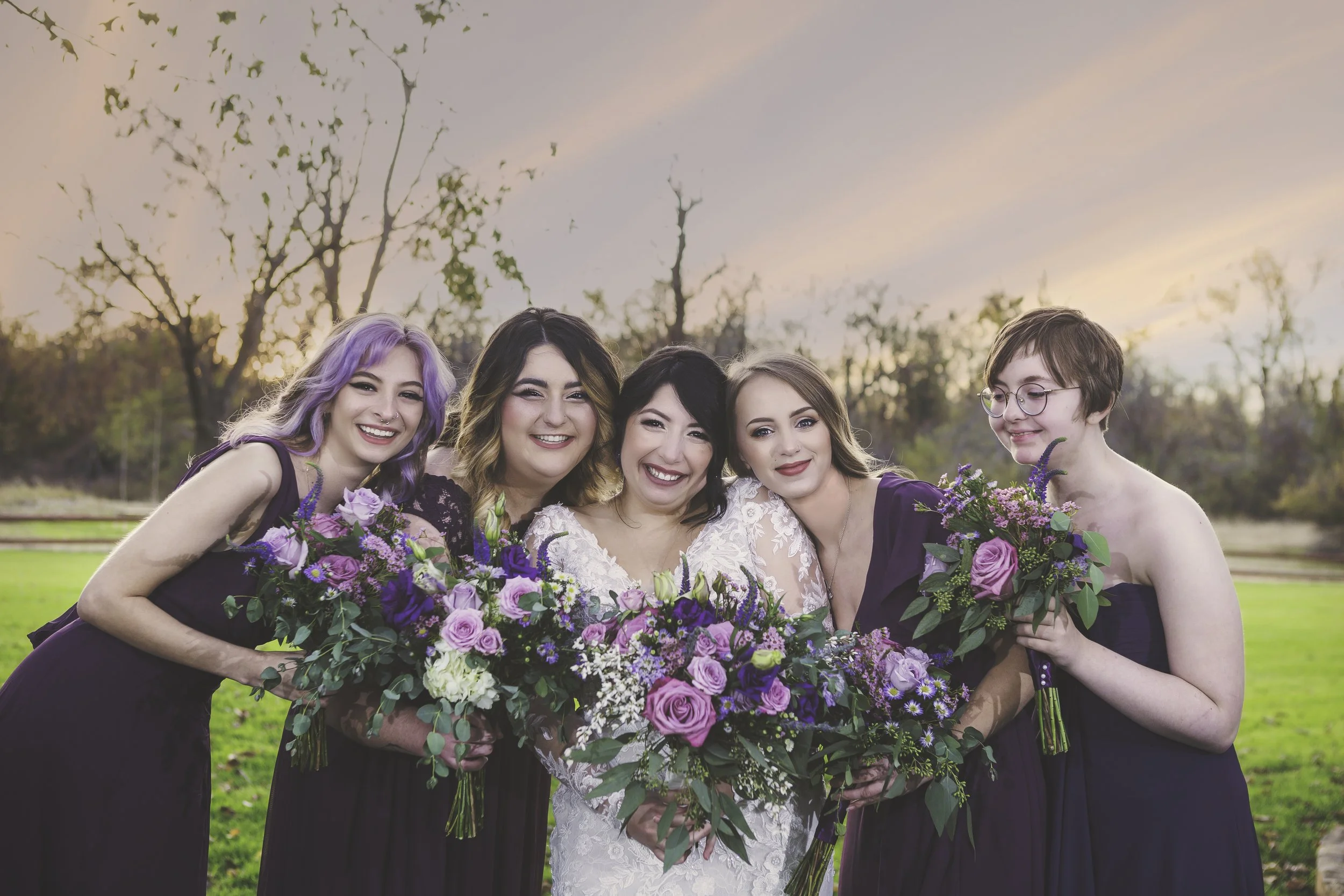 Group of four women in purple dresses and one woman in white dress, holding bouquets of pink and purple flowers, smiling outdoors at sunset