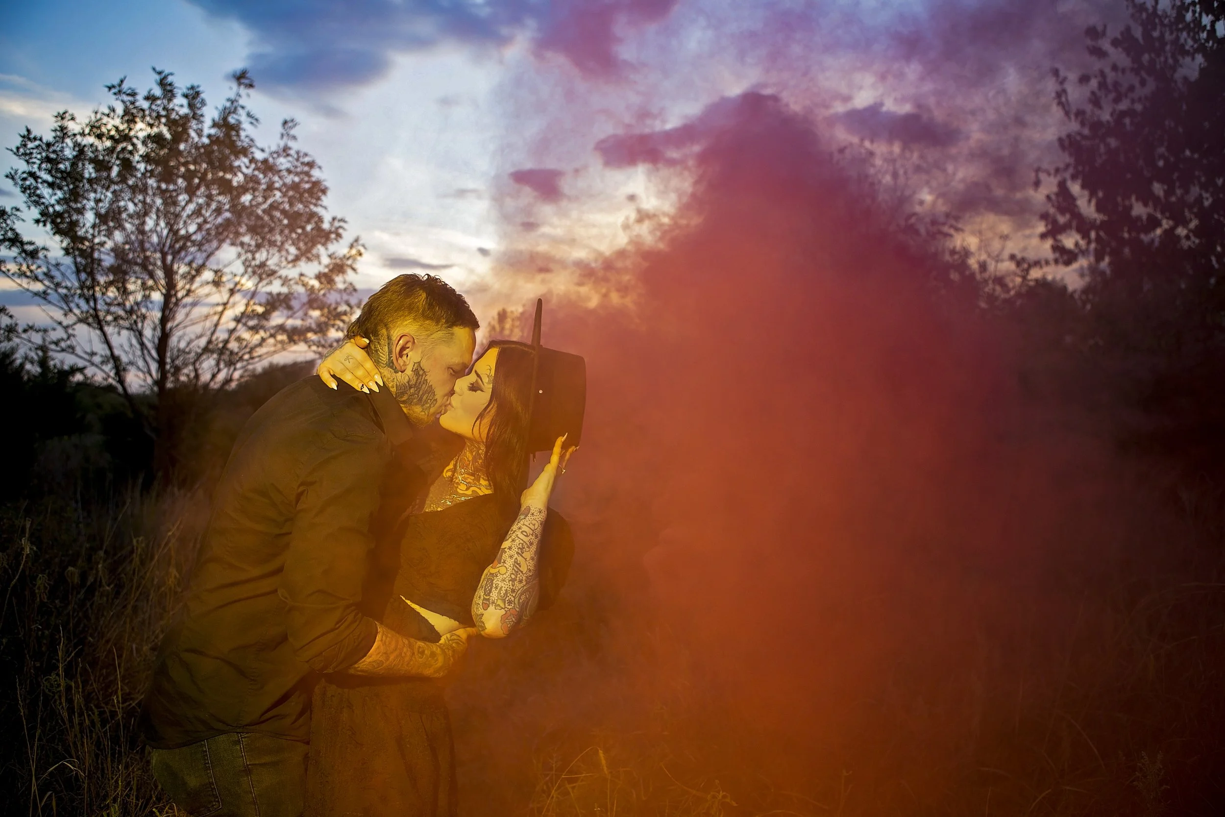 Alternative engagement session with smoke bomb outdoors at sunset, with colorful clouds in the sky and trees in the background.