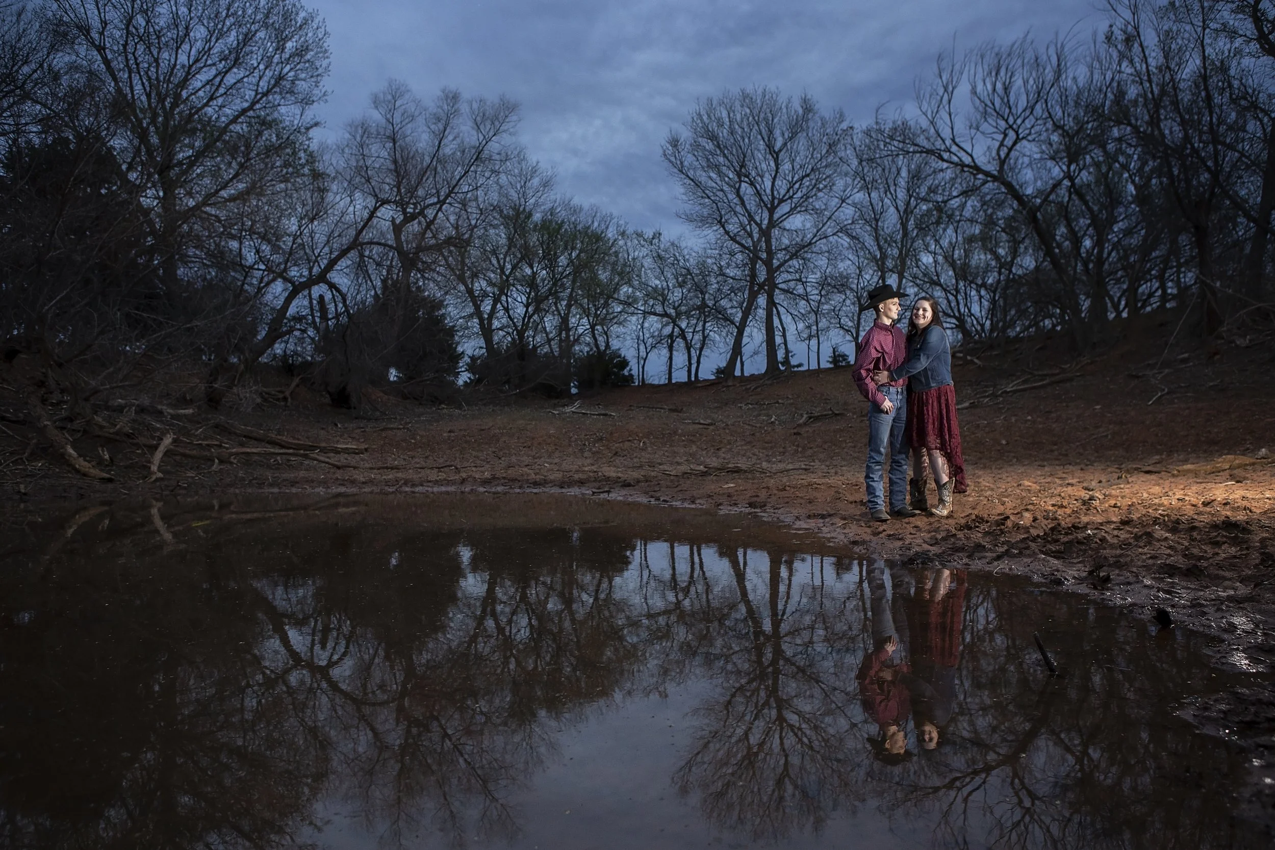 Two women stand close together at the edge of a small pond in a wooded area during dusk, with no leaves on the trees, and their reflection visible in the water.