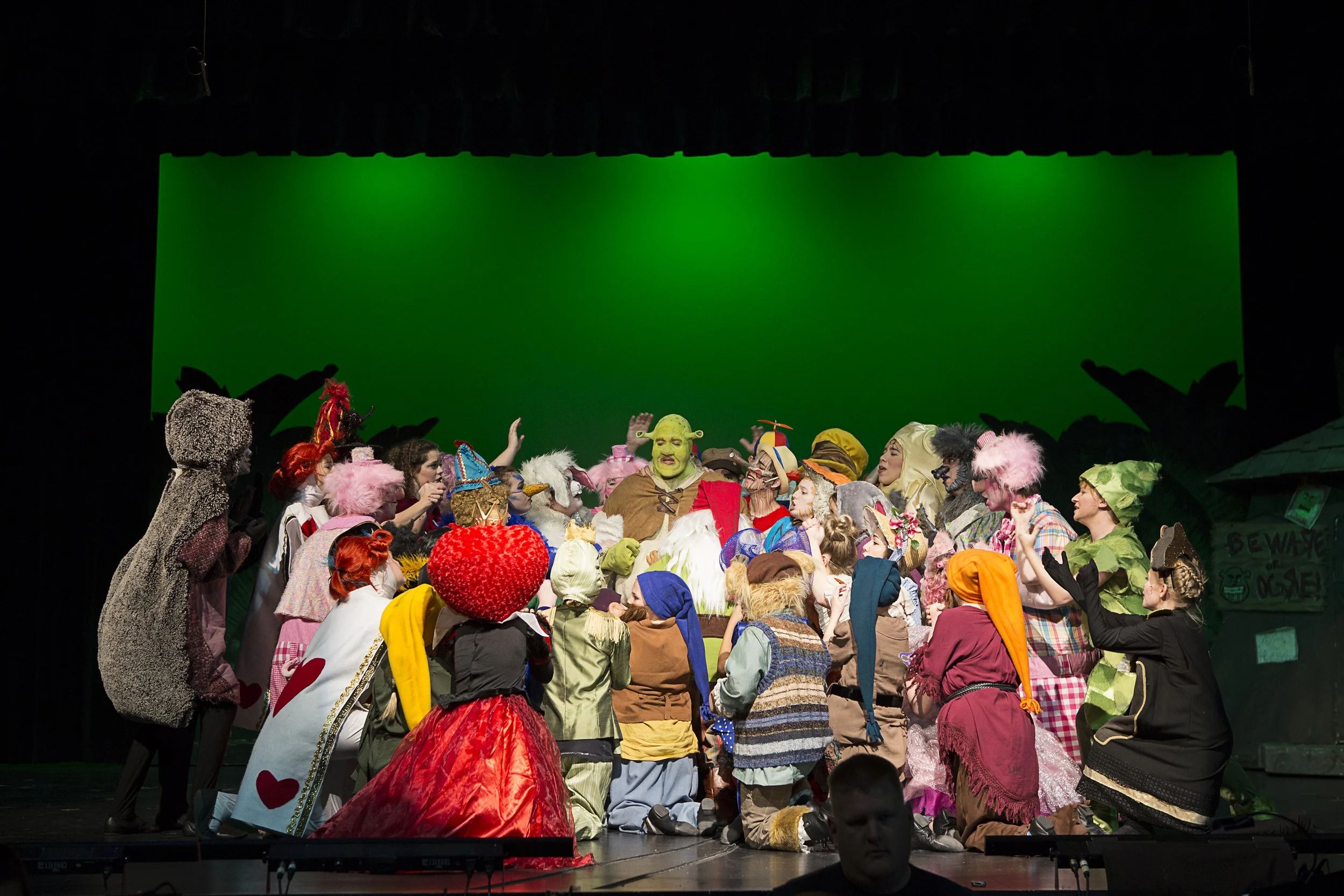 Theater stage with actors in colorful costumes performing in a play against a green screen backdrop.