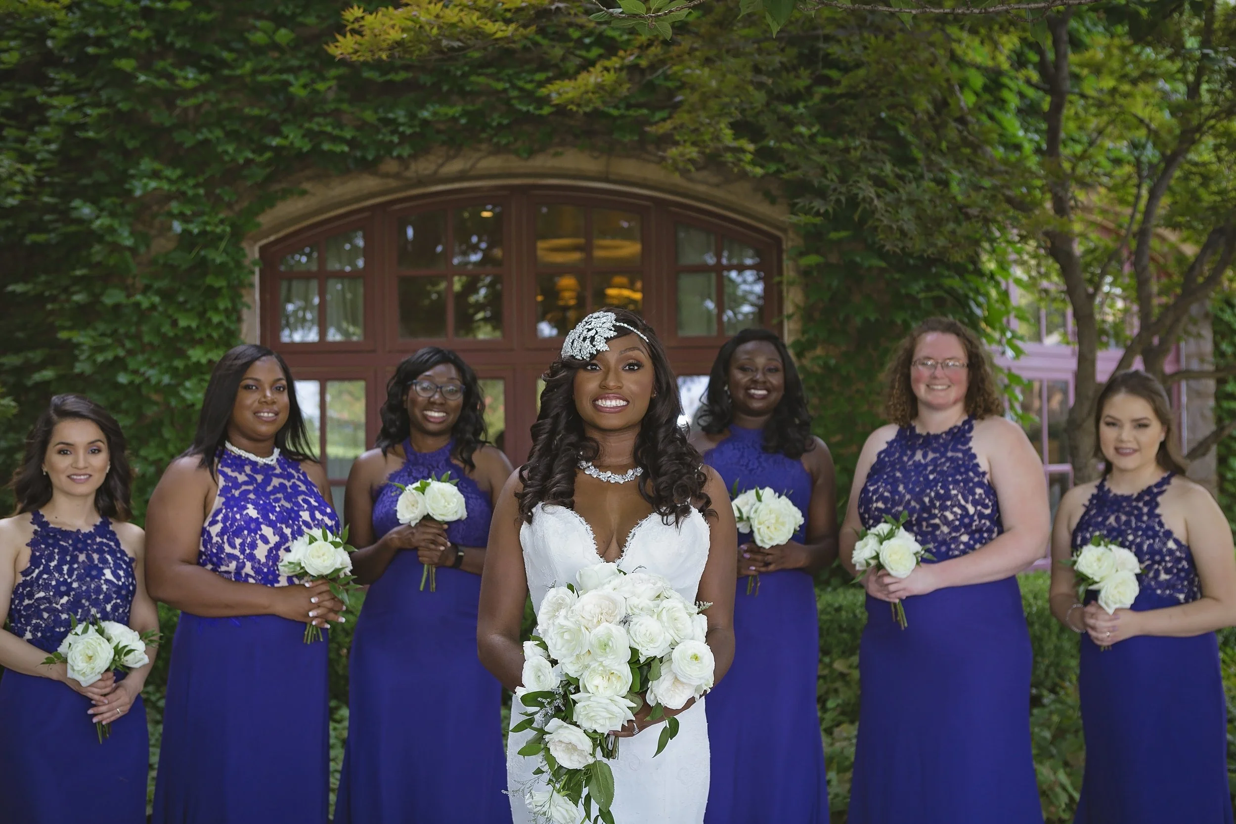 A bride wearing a white dress and a sparkling headpiece is smiling and holding a bouquet of white flowers. She is surrounded by six bridesmaids in matching royal blue dresses with lace tops, all holding white flower bouquets. The group is outdoors in
