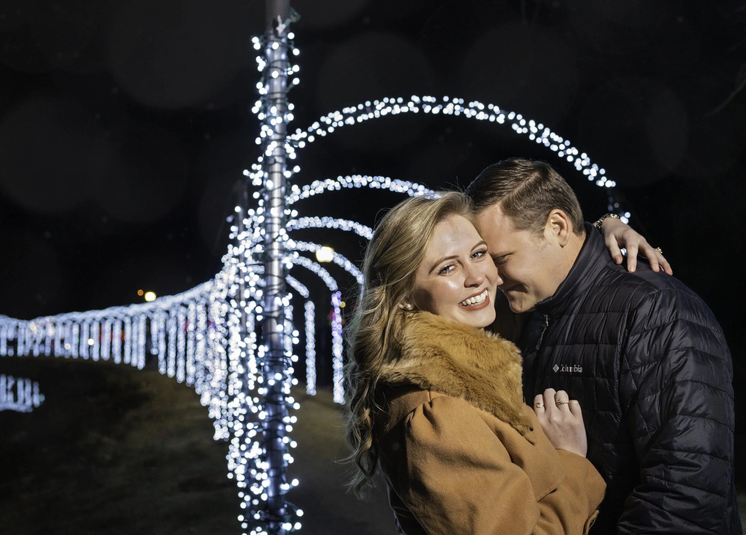 Couple portrait hugging at night in front of Christmas light decorations along a pathway, with the woman smiling and the man whispering into her ear.