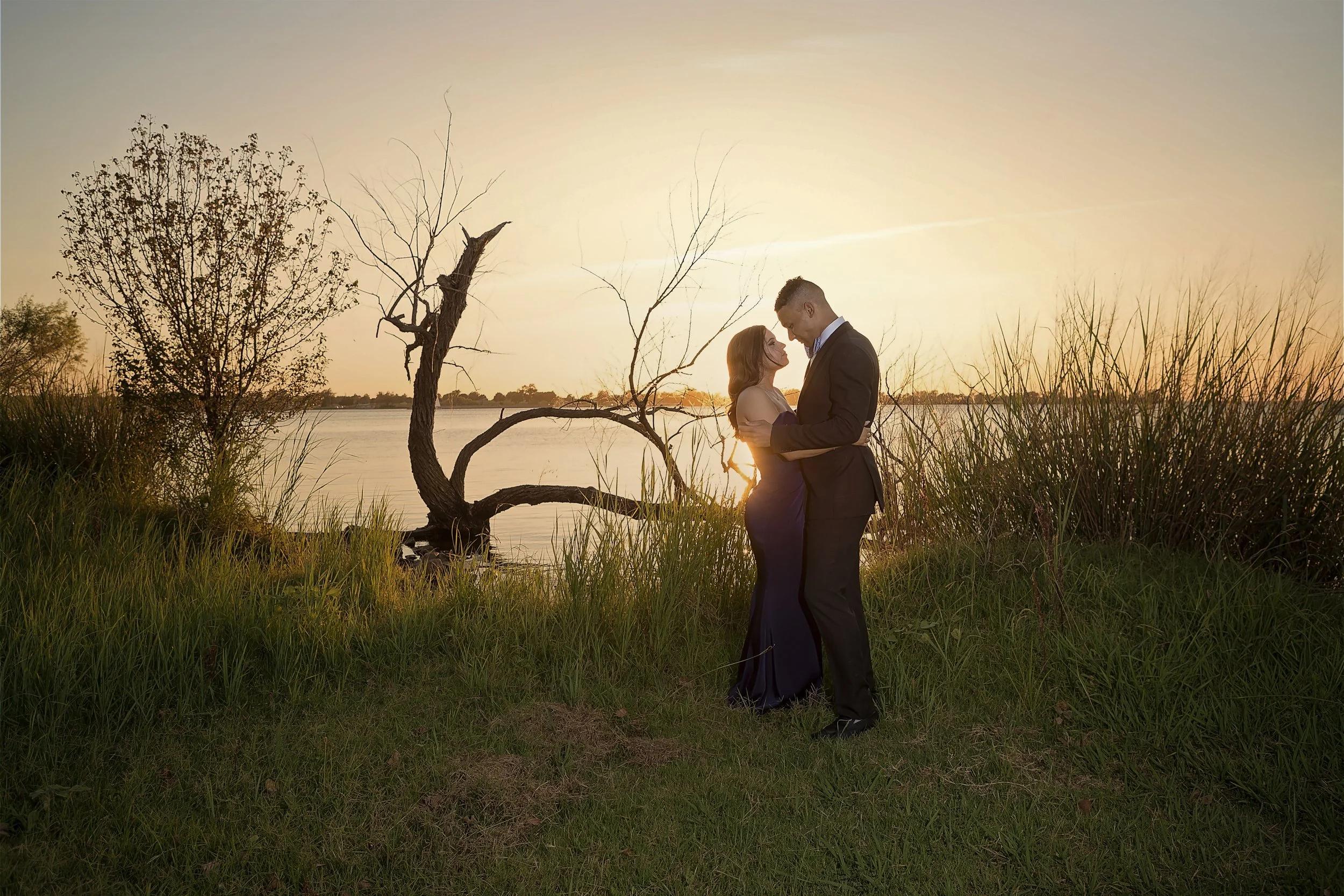 Formal attire engagement session following a planned wedding proposal embracing by a lakeside beach at sunset, with trees and tall grasses surrounding them.