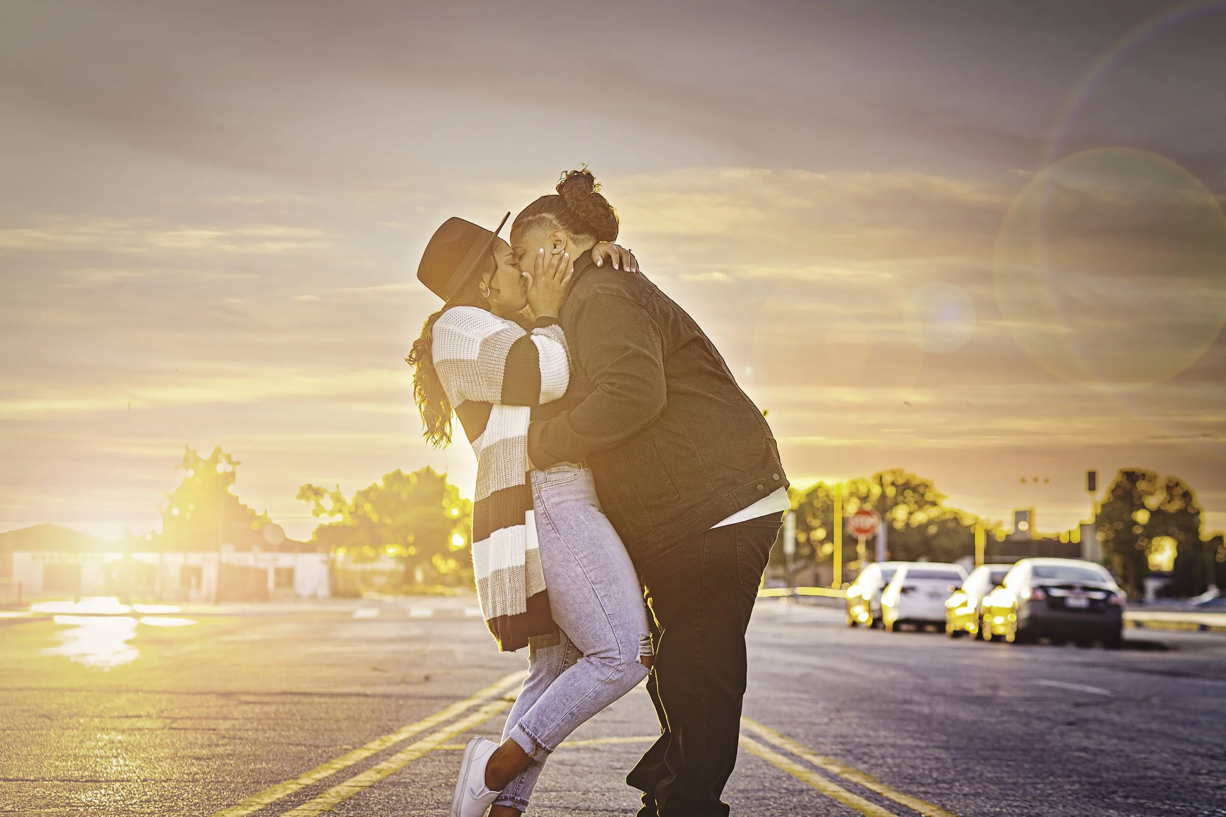 Couple session of sharing a kiss on an empty street during sunset, with cars parked in the background and a rainbow lens flare in the sky.