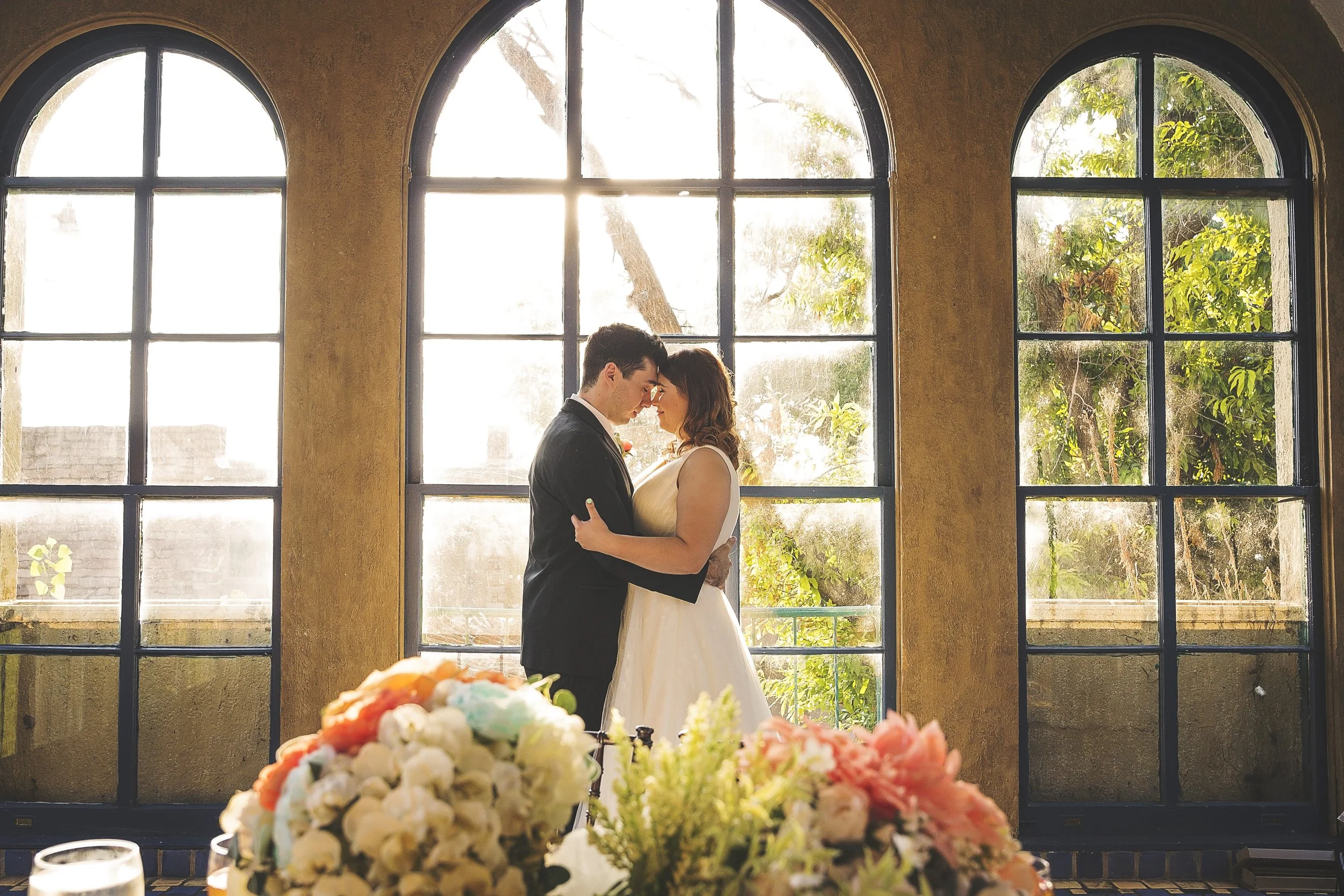 A bride and groom standing close together inside a room with large arched windows, backlit by sunlight. There are floral arrangements in the foreground.