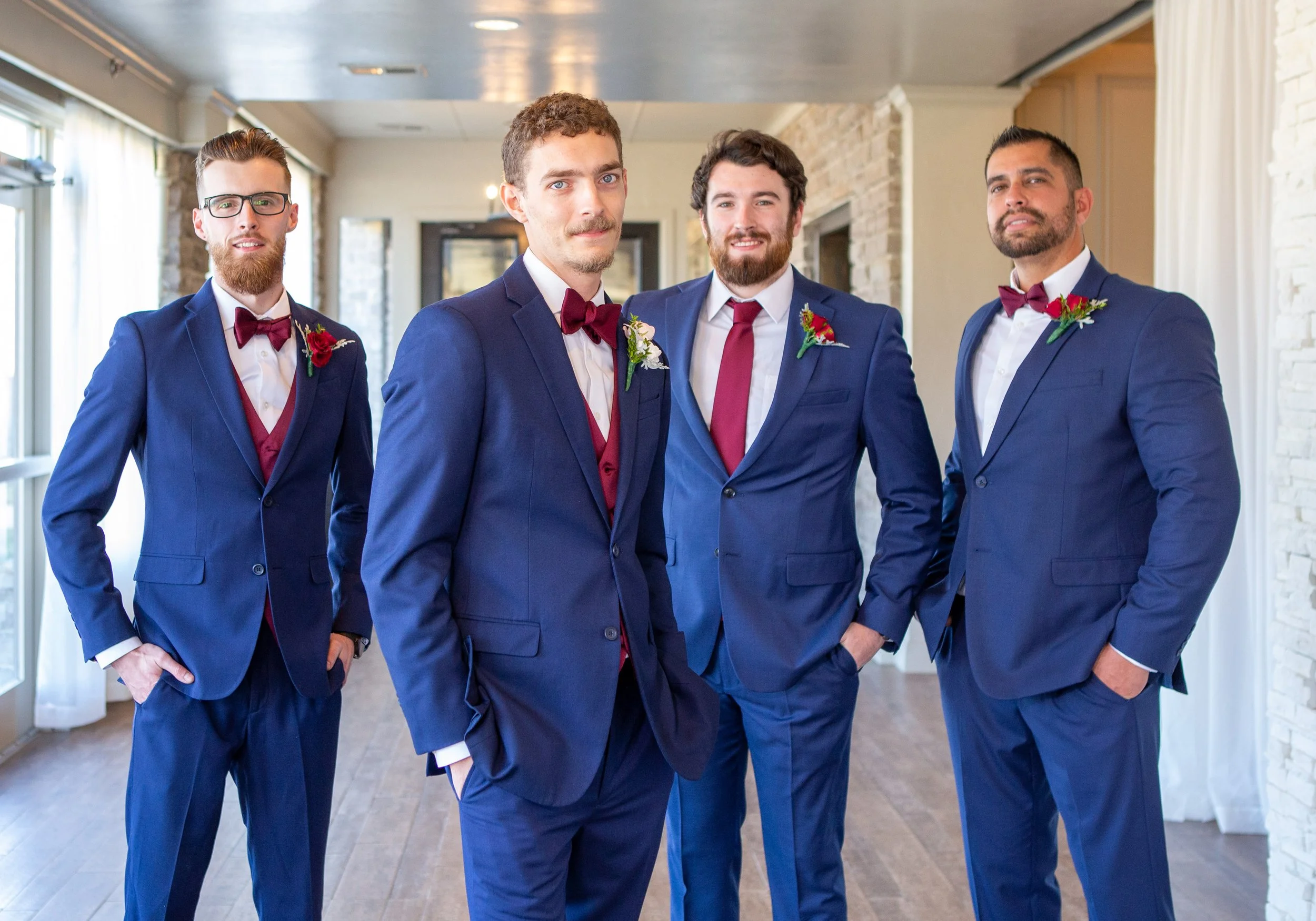 Four men dressed in matching blue suits with white shirts, burgundy bow ties, and boutonnières, standing in a well-lit indoor setting with wood floors and a white brick wall.