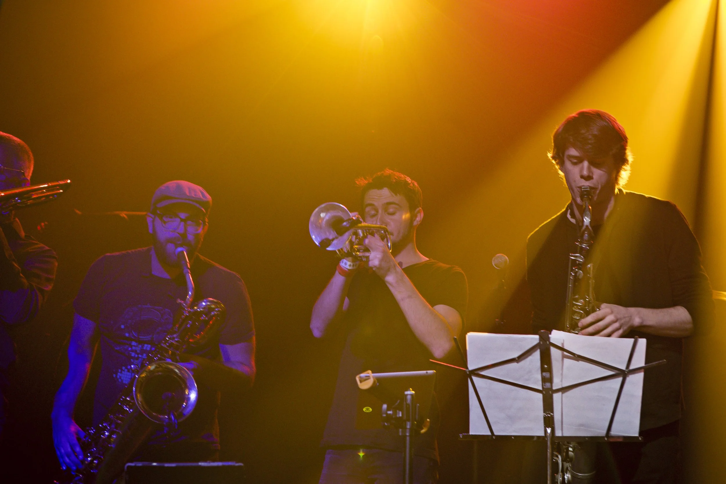 Three musicians on stage playing wind instruments, with warm stage lighting creating a yellow-orange glow.
