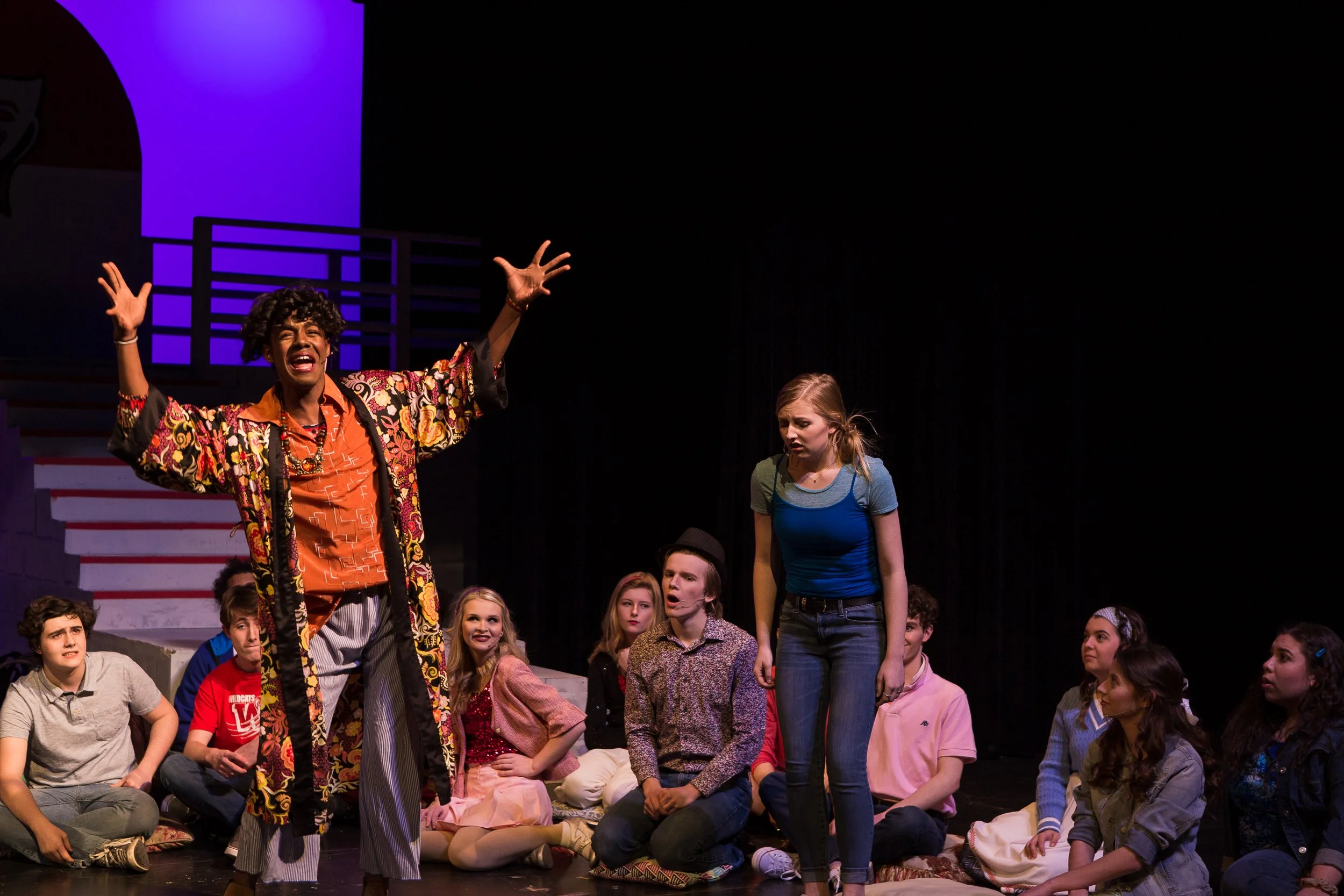 A theatrical performance with a diverse group of young actors, with one person standing and expressive, while others sit and watch, on a stage with dark background and colorful lighting.