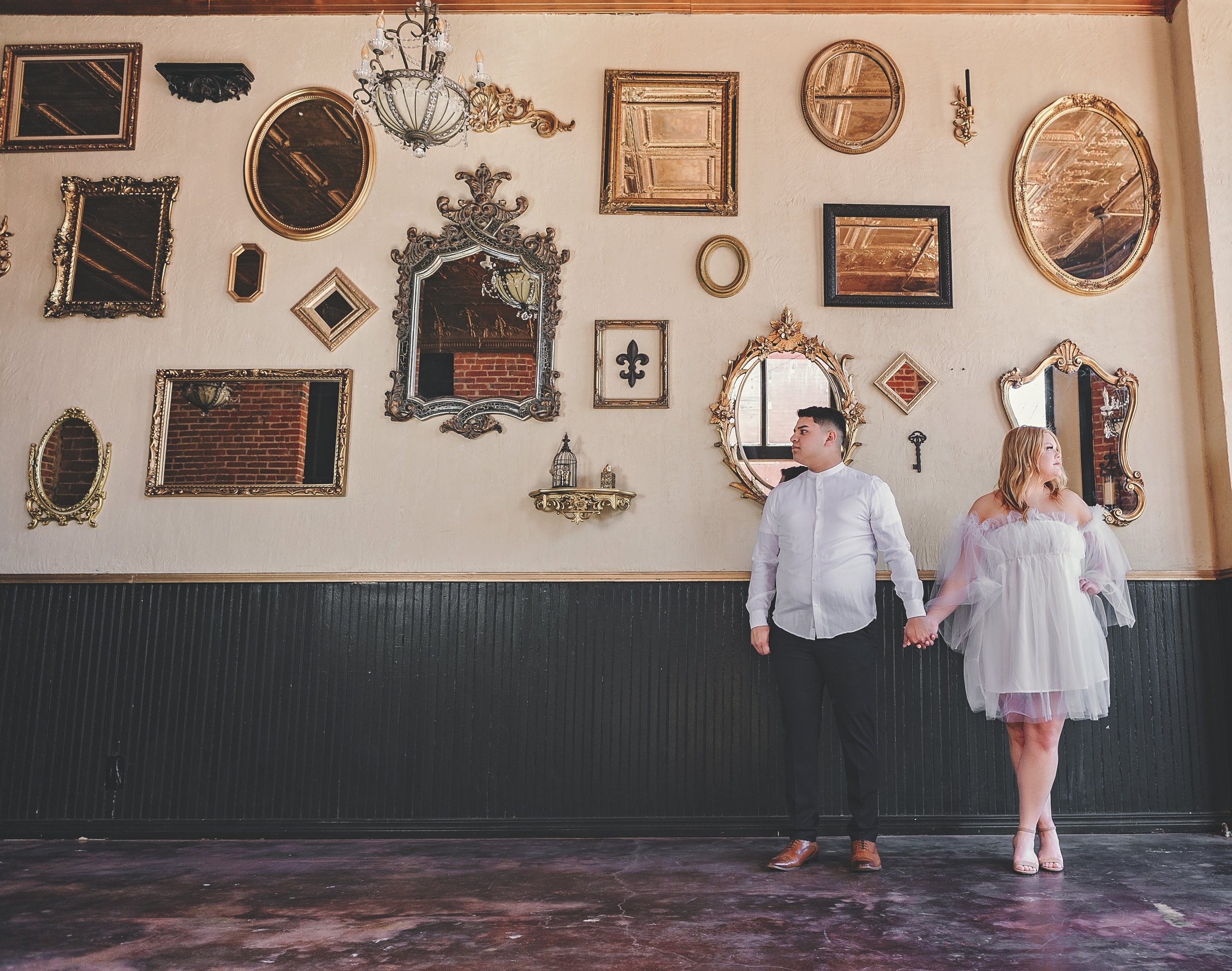 A man and woman holding hands, standing apart against a decorated wall with antique mirrors and picture frames, in a vintage-style interior.