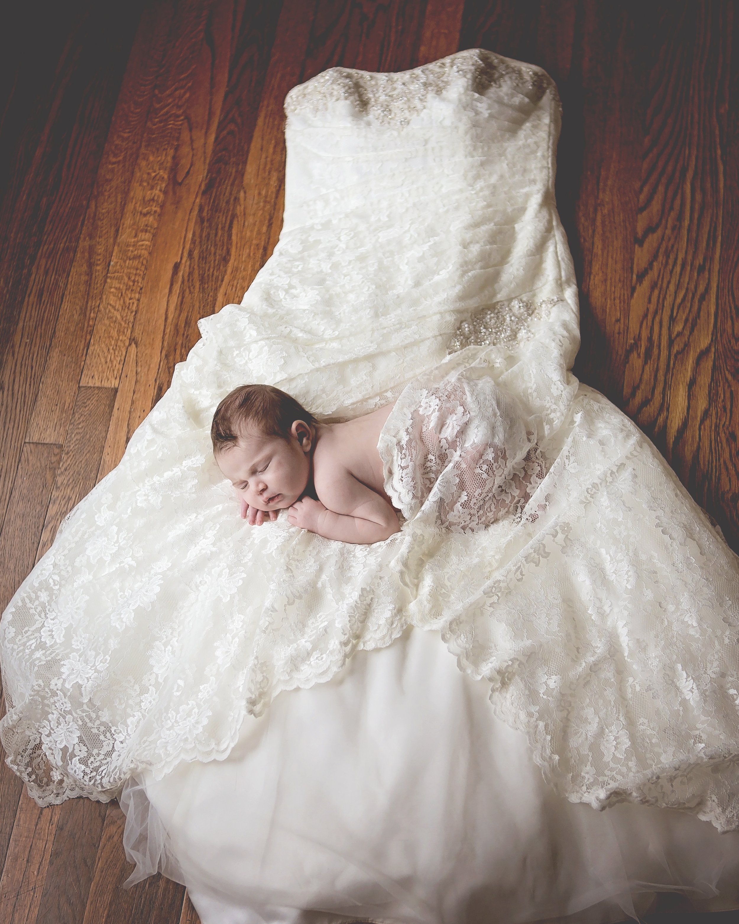 A newborn baby sleeping on a lace and satin wedding dress spread on a wooden floor.