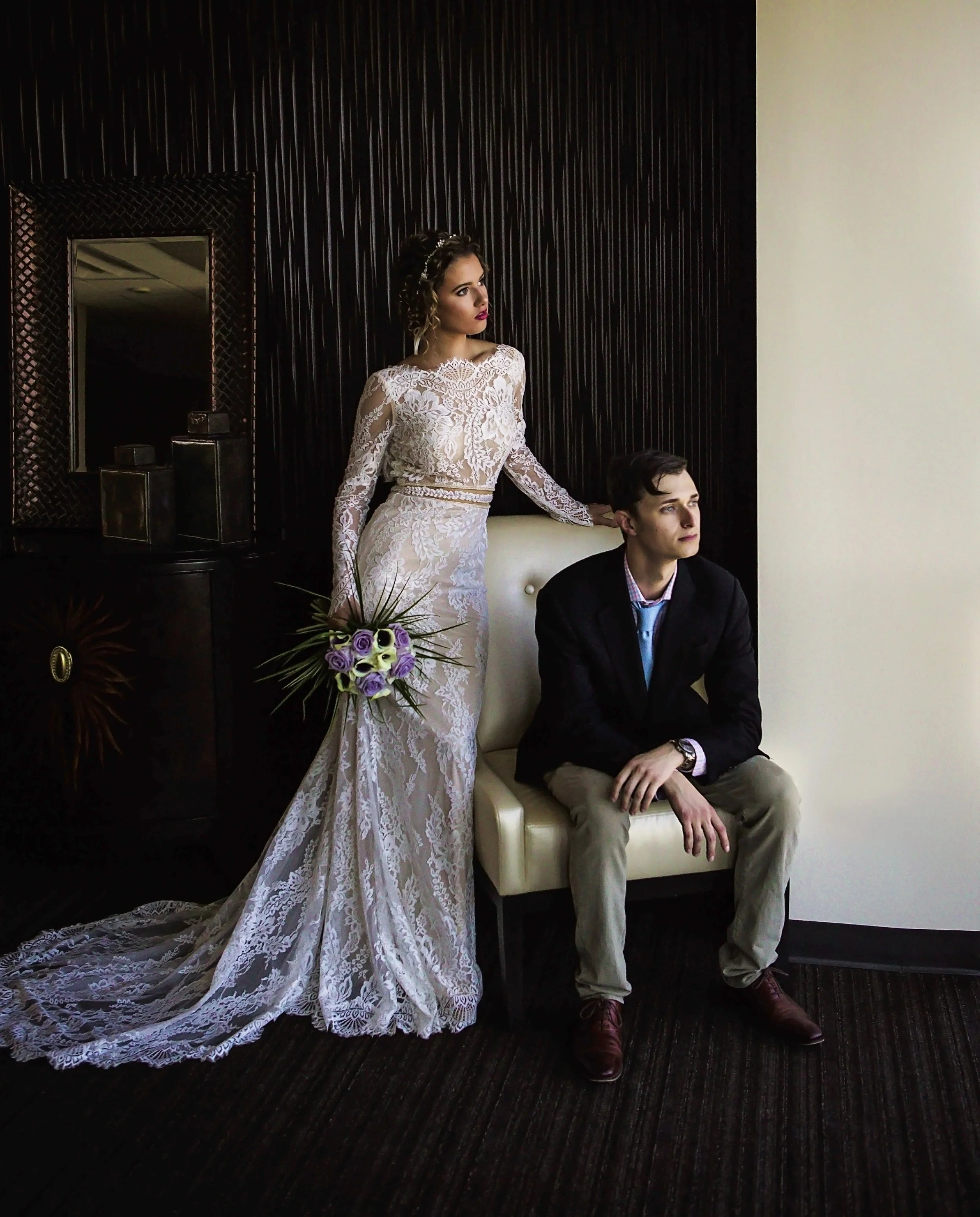 A bride in a white lace wedding dress holding a bouquet, standing next to a seated groom in a dark blazer and beige pants, in an indoor setting with dark wood paneling and a cream wall.