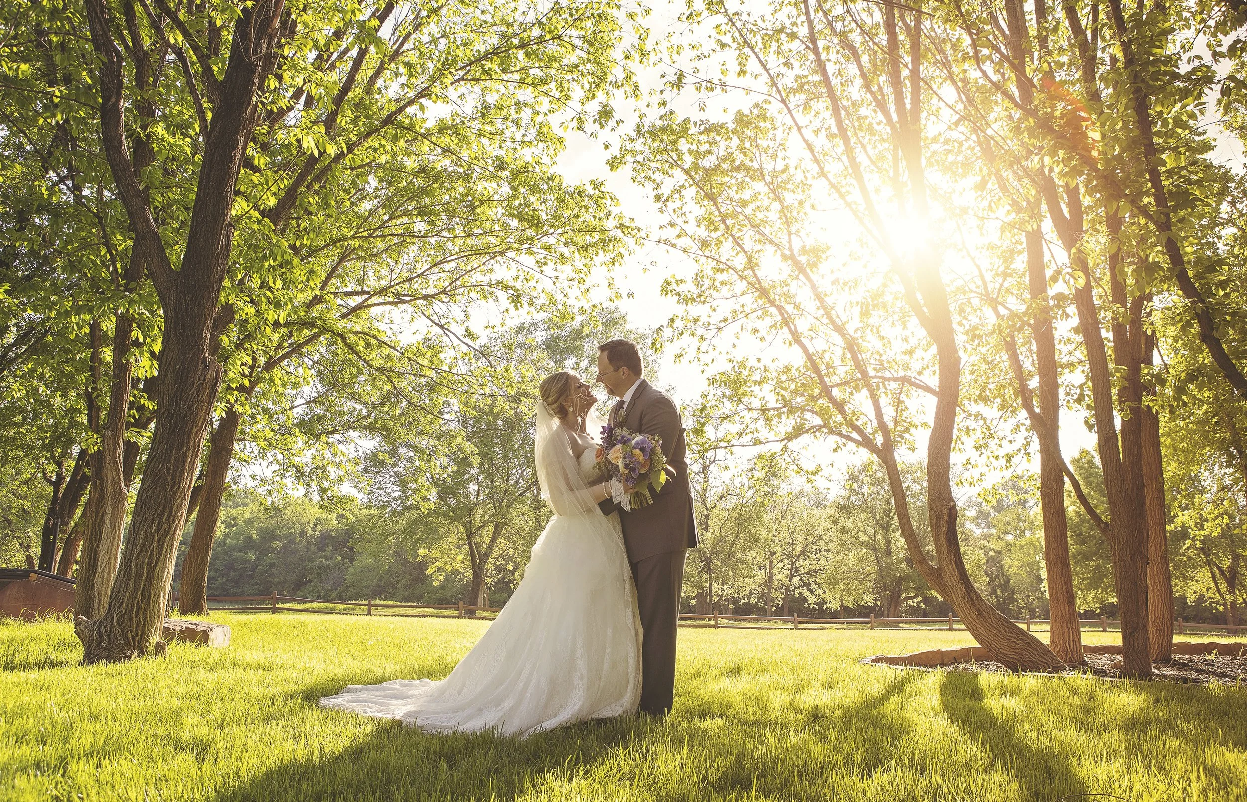 A bride and groom standing close together in a sunlit park with green trees and grass, the bride holding a bouquet of flowers, during a wedding ceremony or photo shoot.