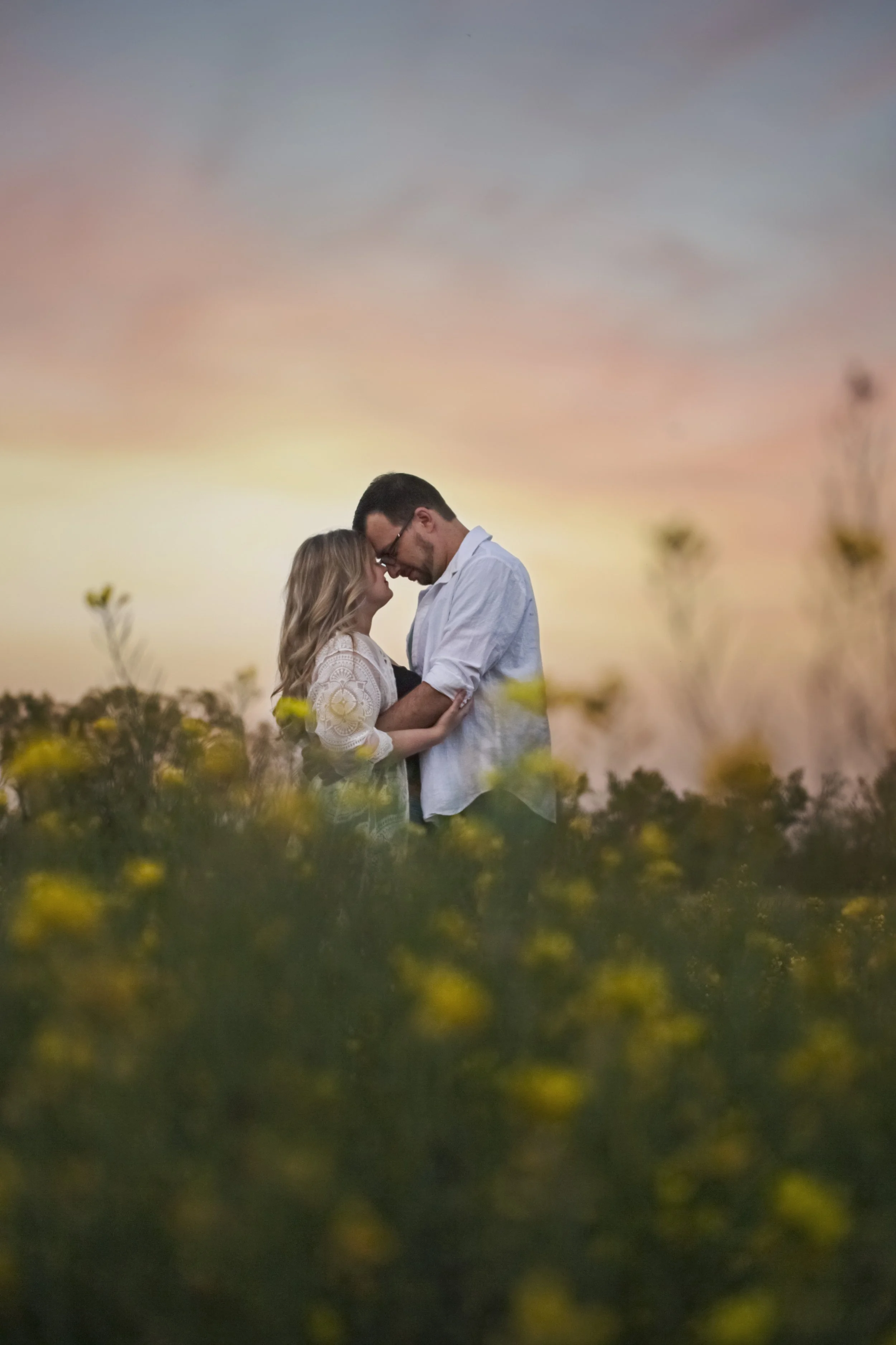 Engagement session of a couple stands close together in a field of yellow flowers during sunset, with their foreheads touching and eyes closed.