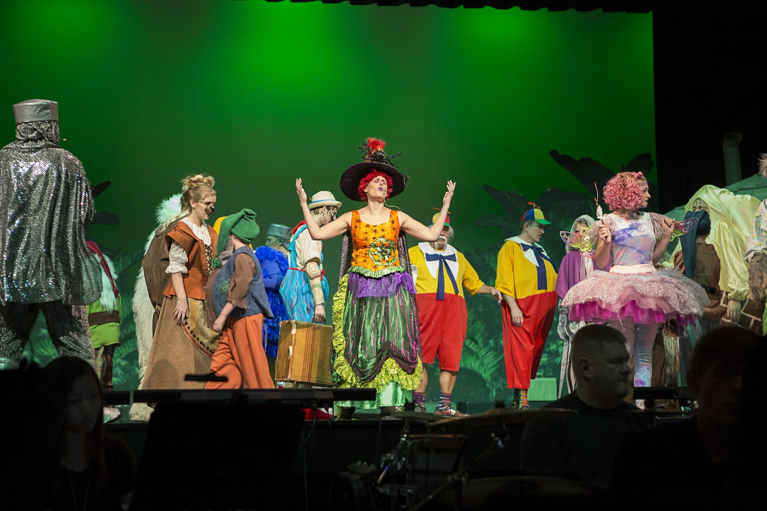 A theatrical stage scene with actors in colorful costumes, including a woman in a large purple and green dress with a wide-brimmed hat, standing in the center with arms raised. Other performers wear bright, whimsical clothing and accessories, set aga
