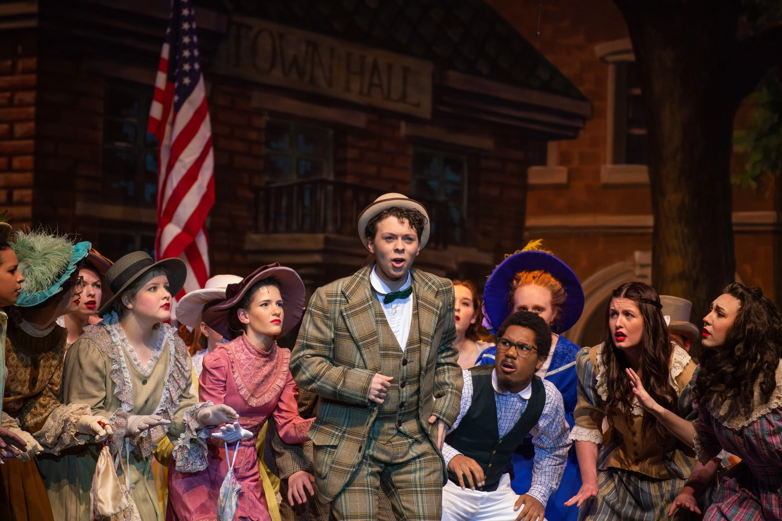A group of actors performing on stage, dressed in vintage clothing, with one person in the center wearing a plaid suit and a hat, appearing to sing or speak passionately. An American flag is visible in the background.