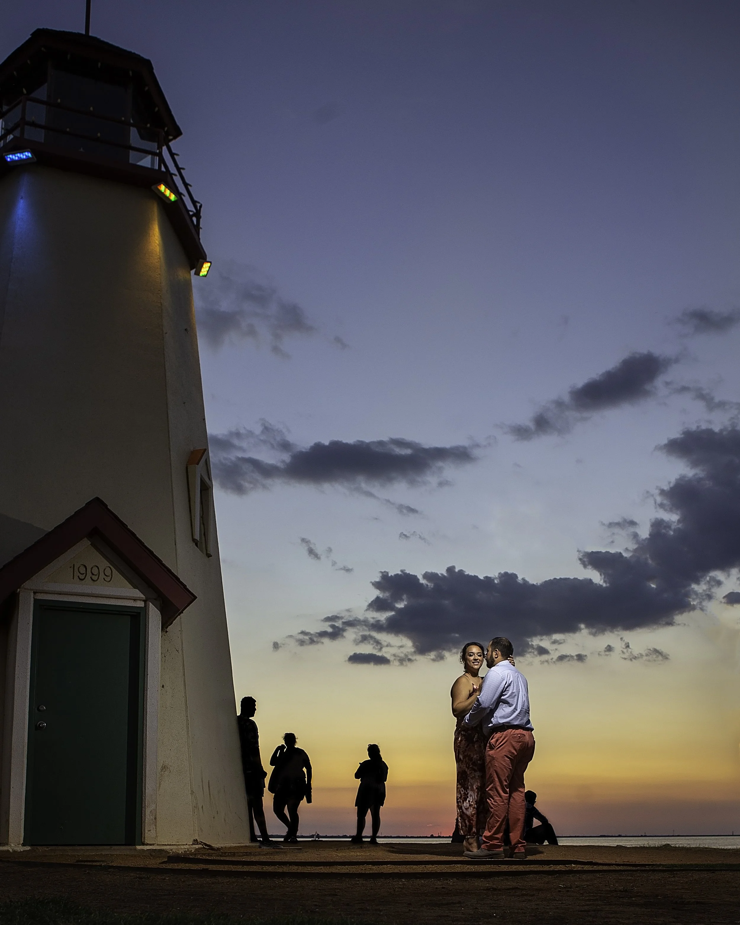 A lighthouse, with a group of people under a sunset sky featuring orange and purple clouds. Newly engaged couple in the foreground are dancing or talking, while others are standing or sitting near the water.