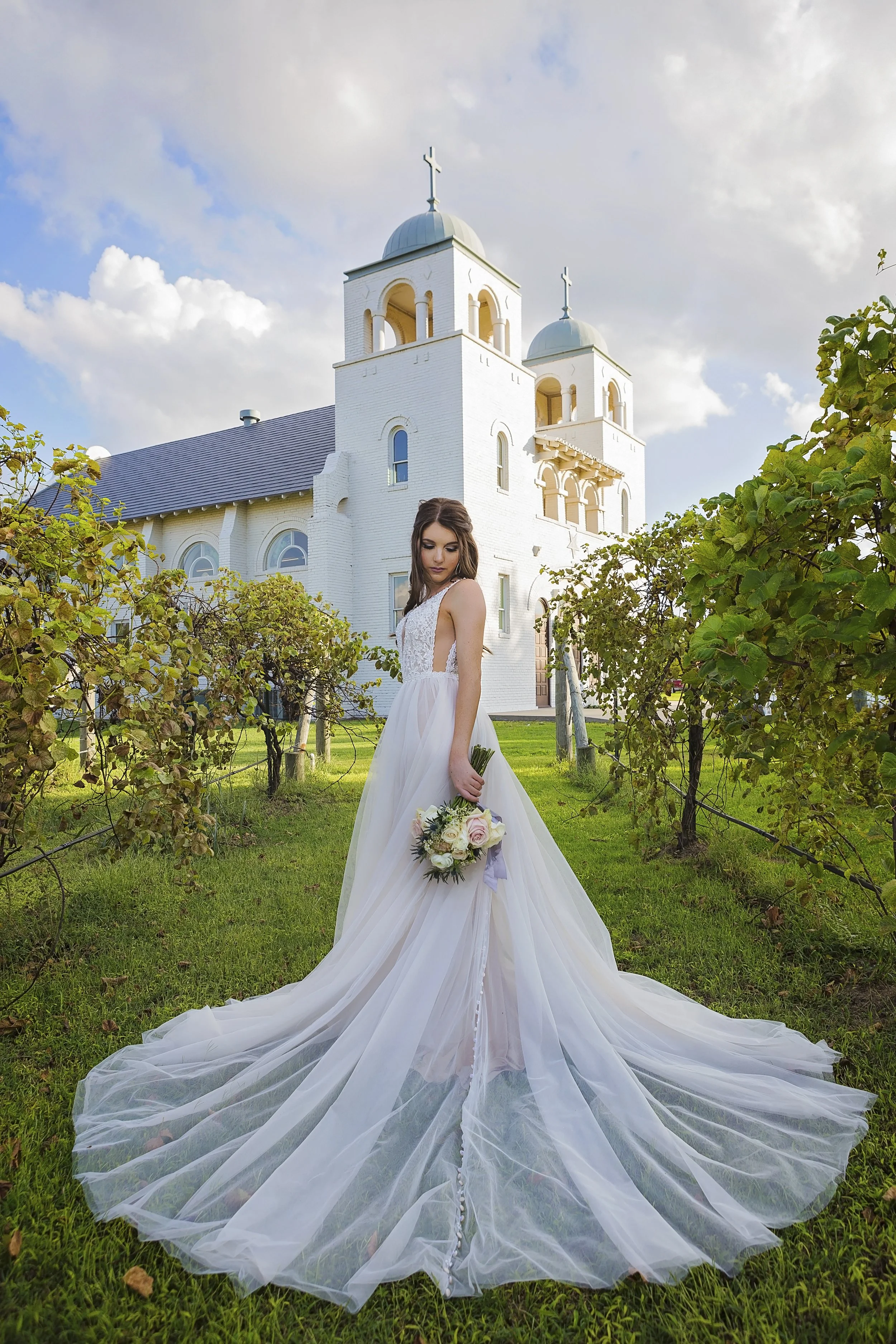 A bride in a white wedding dress holding a bouquet, standing on a grassy path between vineyard rows, with a white church with two domed towers in the background on a partly cloudy day.