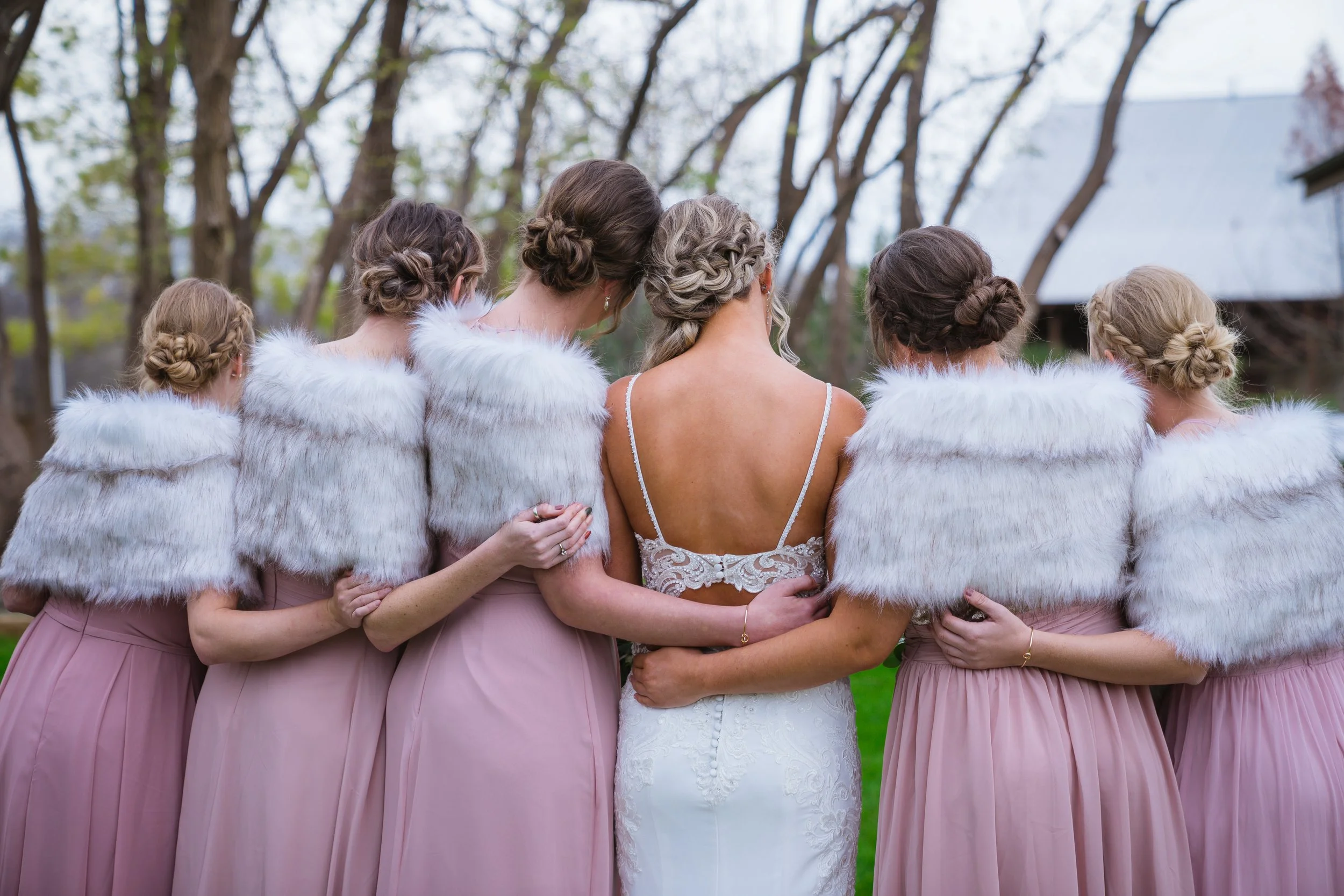 Bridesmaids and bride with braided hairstyles hugging outdoors with trees in the background.