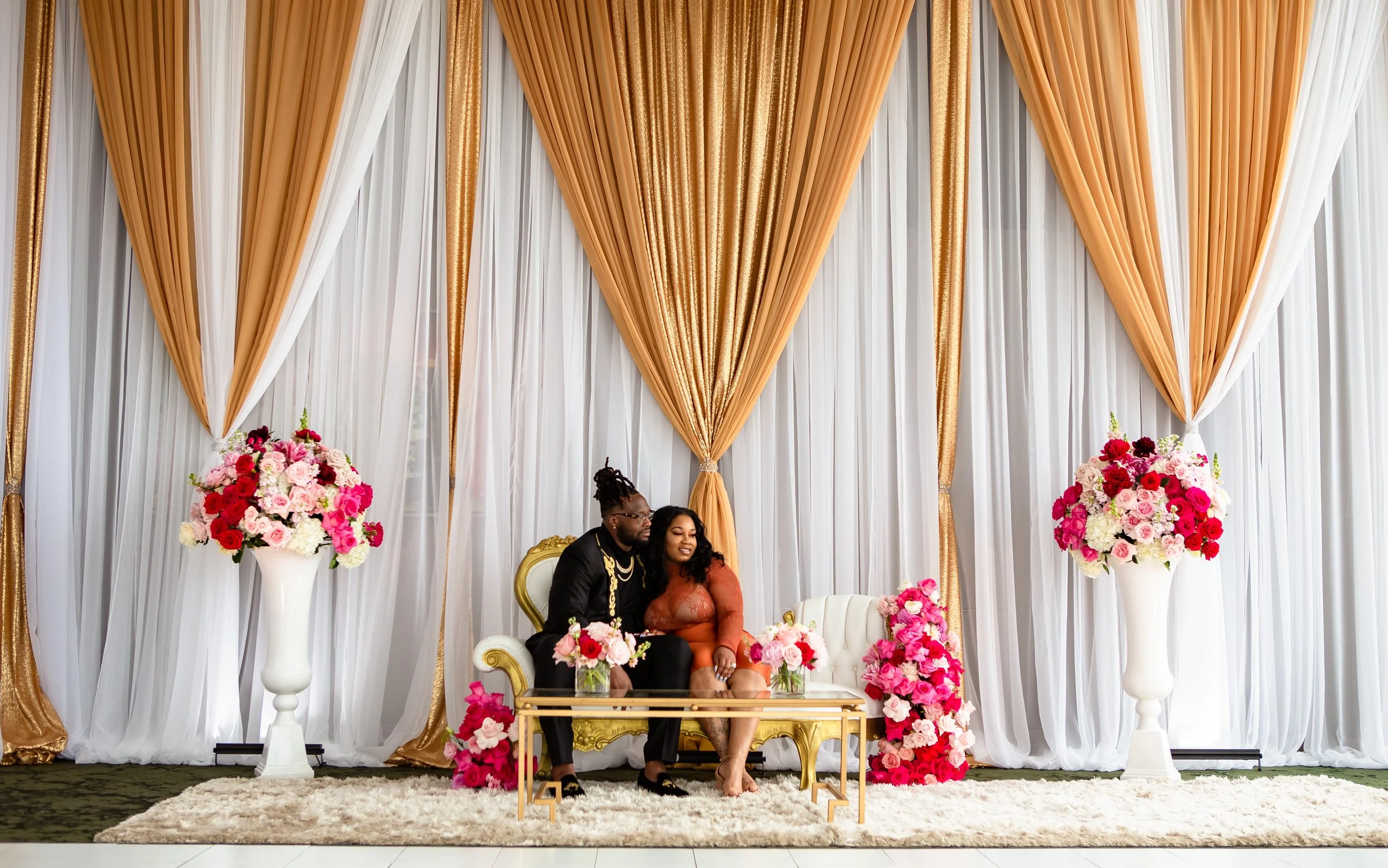 A couple sitting on a white and gold ornate couch, surrounded by large flower arrangements with pink, red, and white flowers, in front of white and gold drapery.