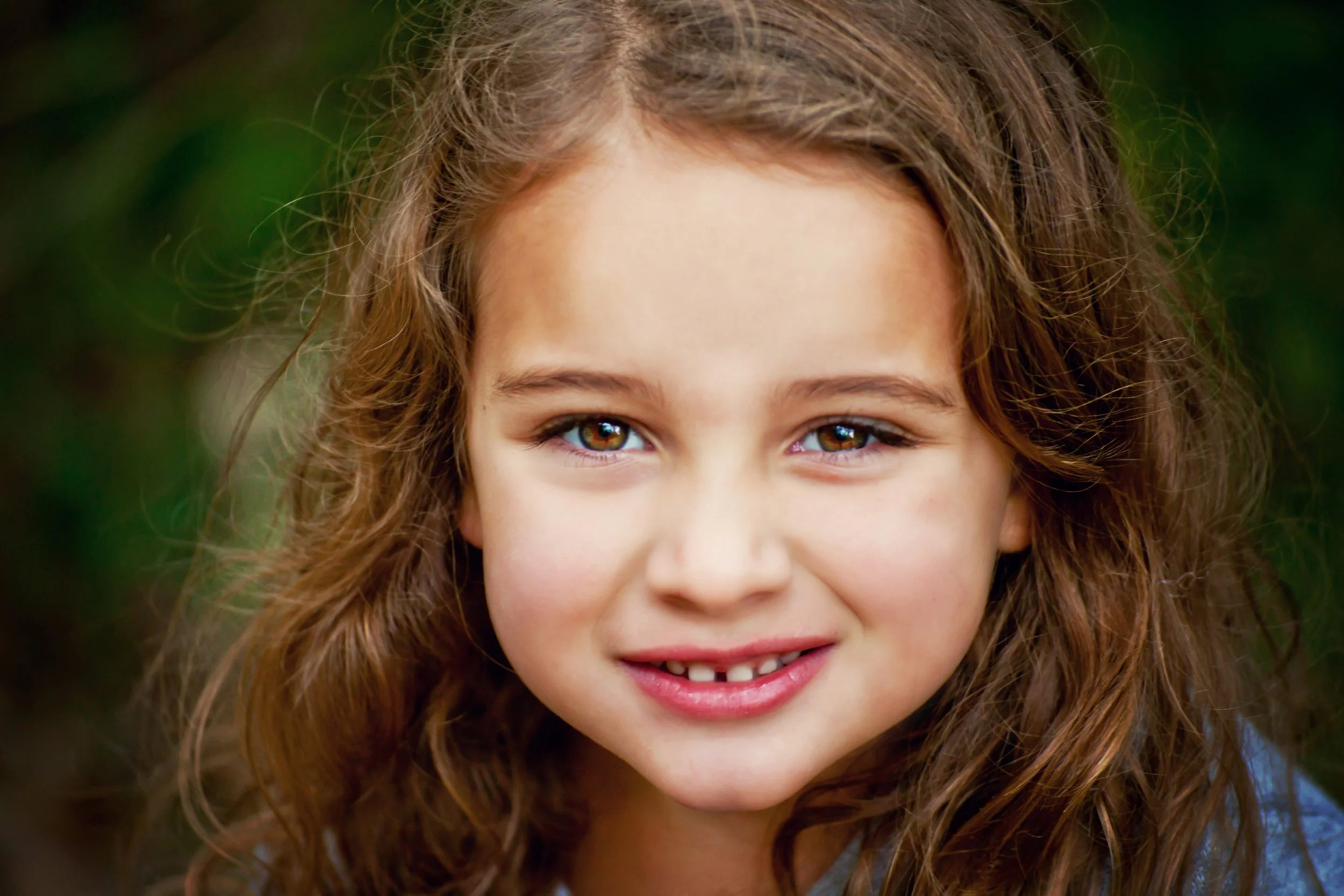 Close-up shot of a young girl with curly brown hair, bright eyes, and a slight smile, outdoors with a blurred natural background.