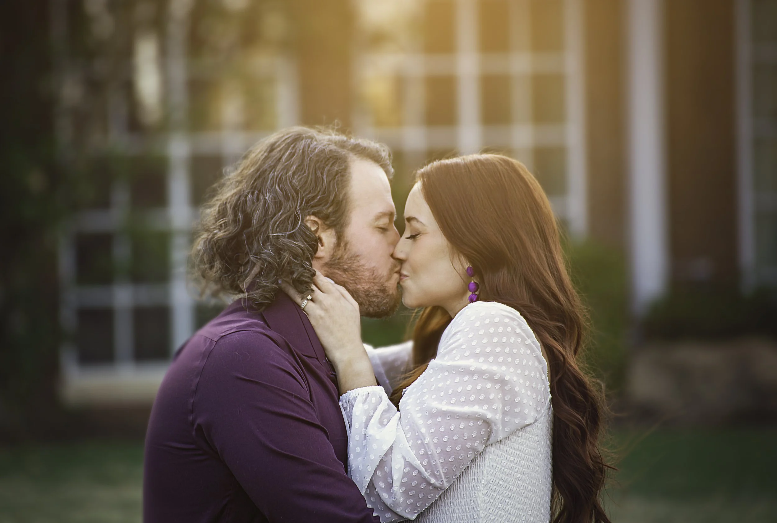 Outdoor engagement session during sunset, with their eyes closed and faces touching, in front of a building with large windows.