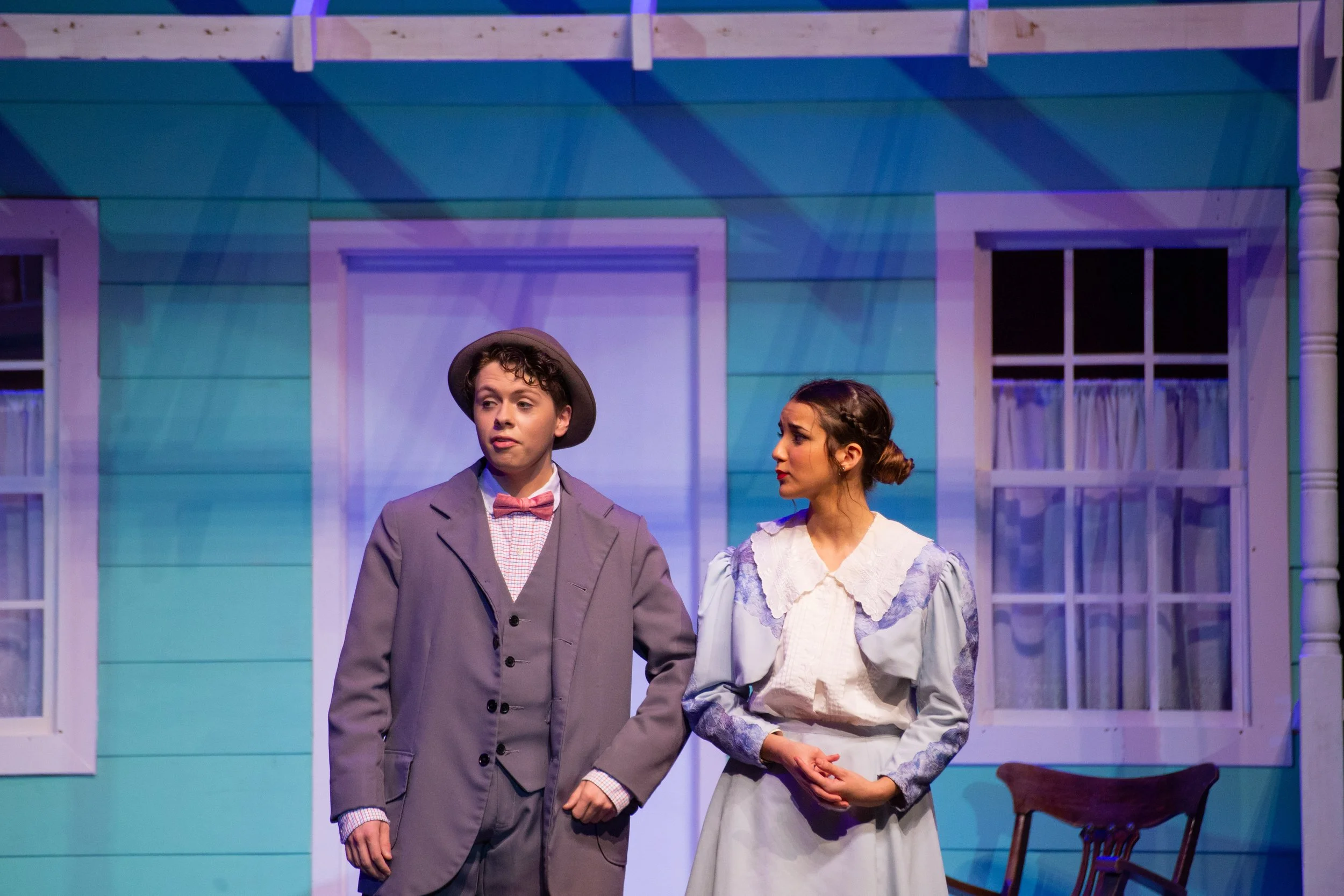 Two actors on stage portraying a scene from a play, with a blue house backdrop, one man in a gray suit and hat, and woman in a light-colored dress.