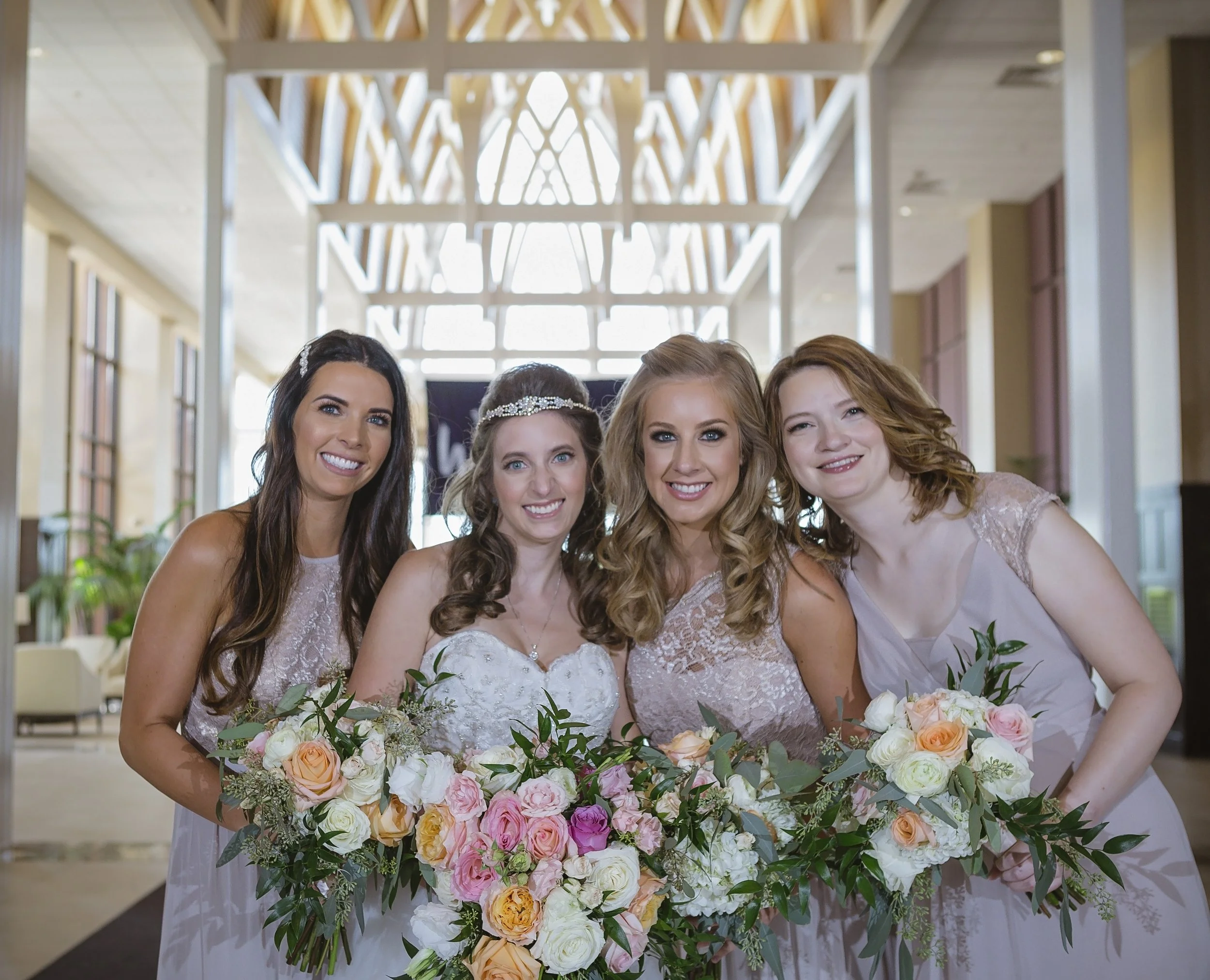 Four women dressed in light-colored dresses standing close together and holding large bouquets of flowers, smiling at the camera in a bright, modern indoor setting.