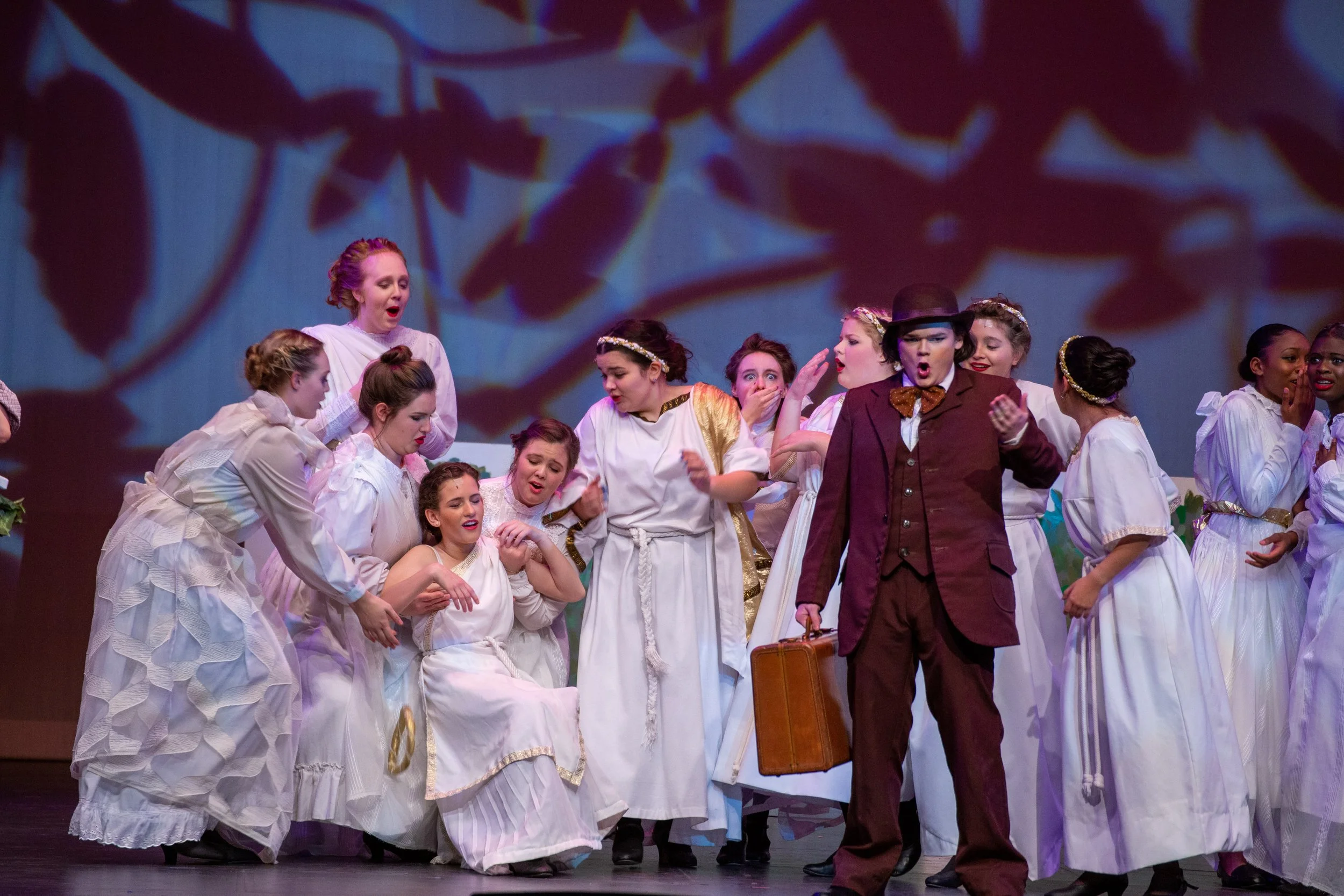 Theatrical scene with multiple women dressed in white, and a man in a suit with a hat and briefcase, on stage with a background projection.