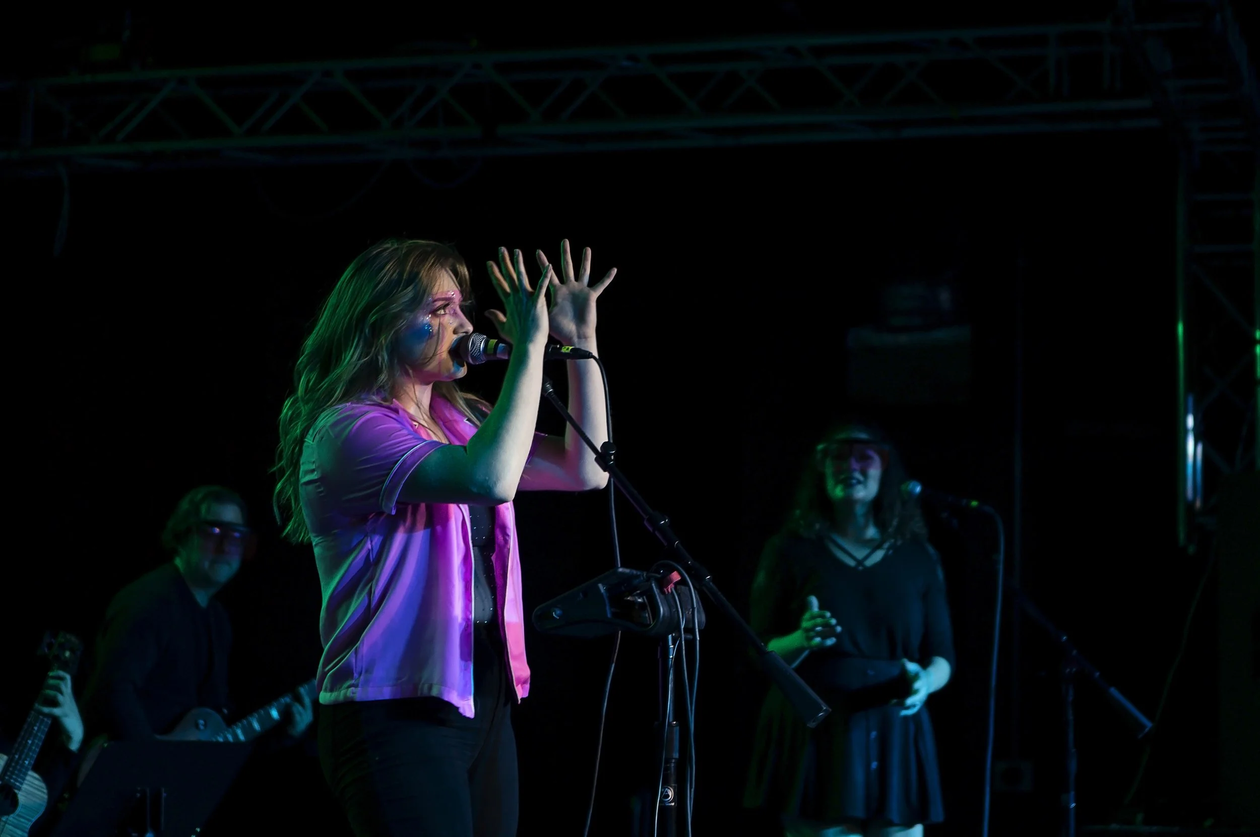 Female singer performing on stage with backup singers and guitarist, with stage lighting and sound equipment.
