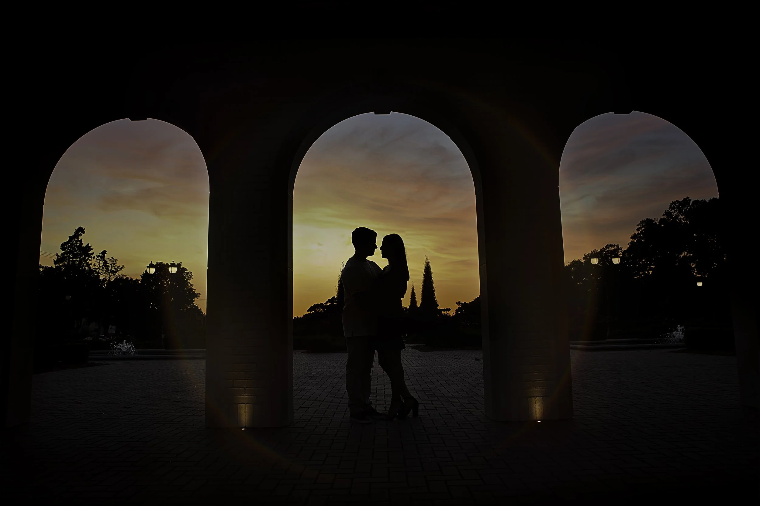 Silhouette of a couple standing close together under an arched structure at sunset, with trees and a fountain in the background.