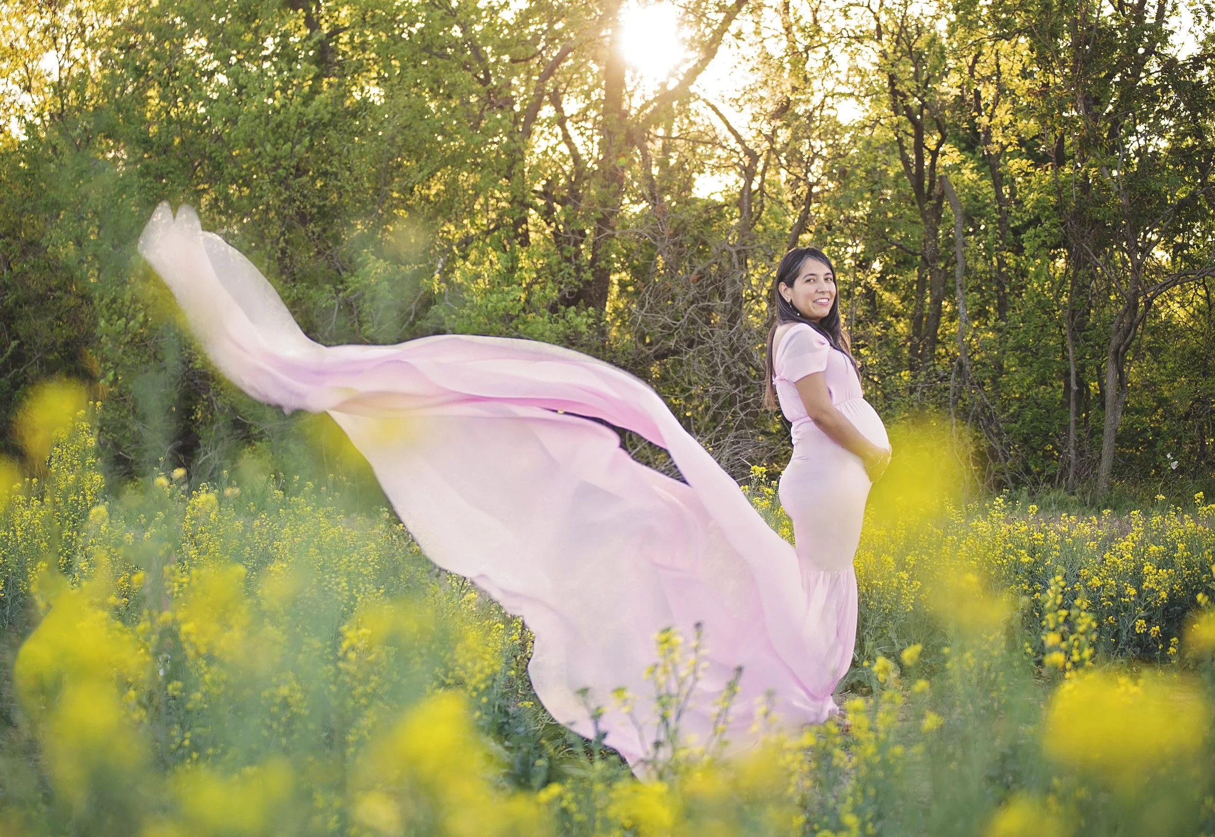Pregnant woman in a flowing pink dress standing in a field of yellow flowers with trees in the background sunlight filtering through the trees.