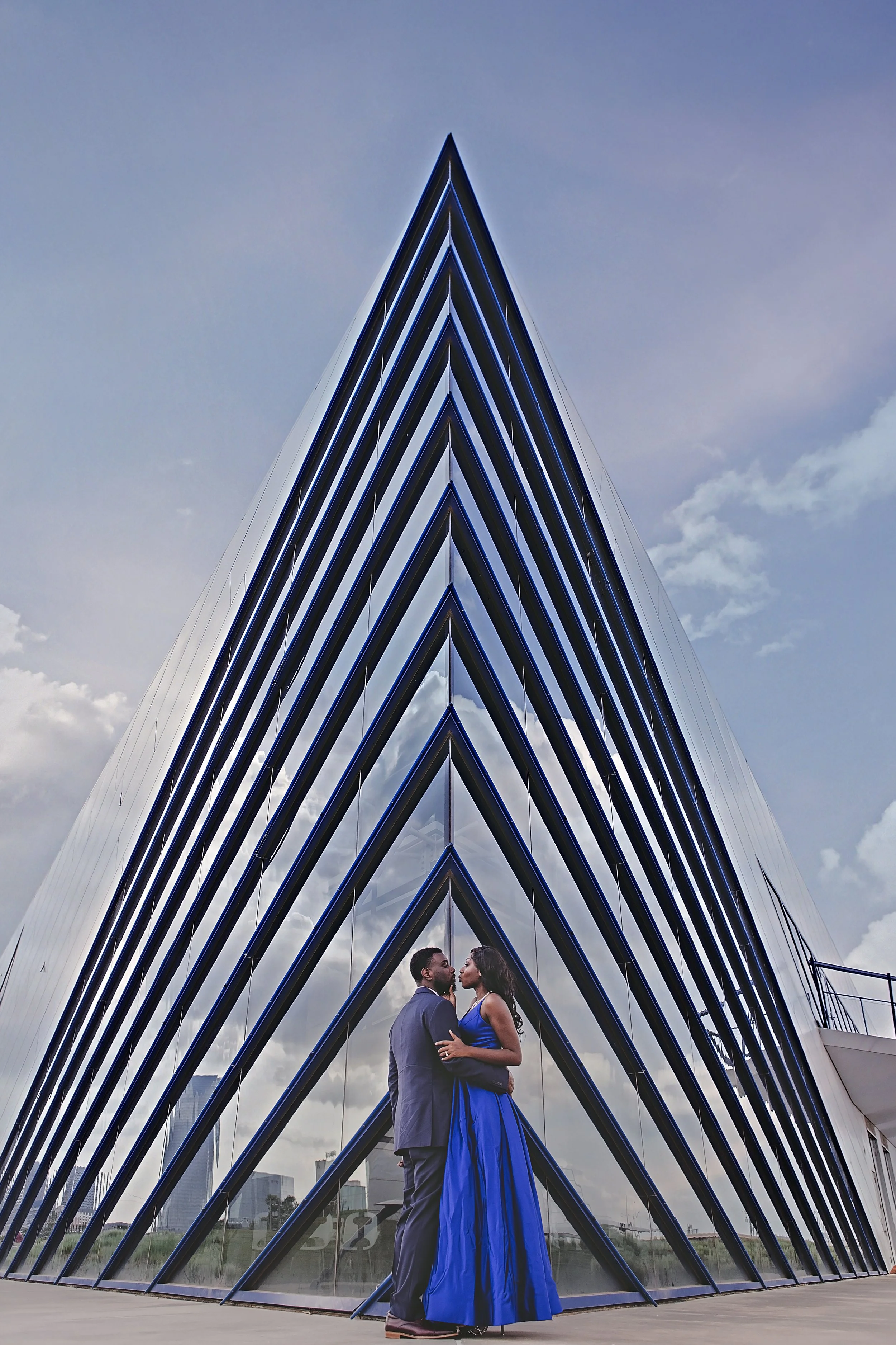 A couple dressed in formal attire standing close together in front of a modern glass and metal pyramid-shaped building with blue and black accents, with city buildings and a cloudy sky in the background.